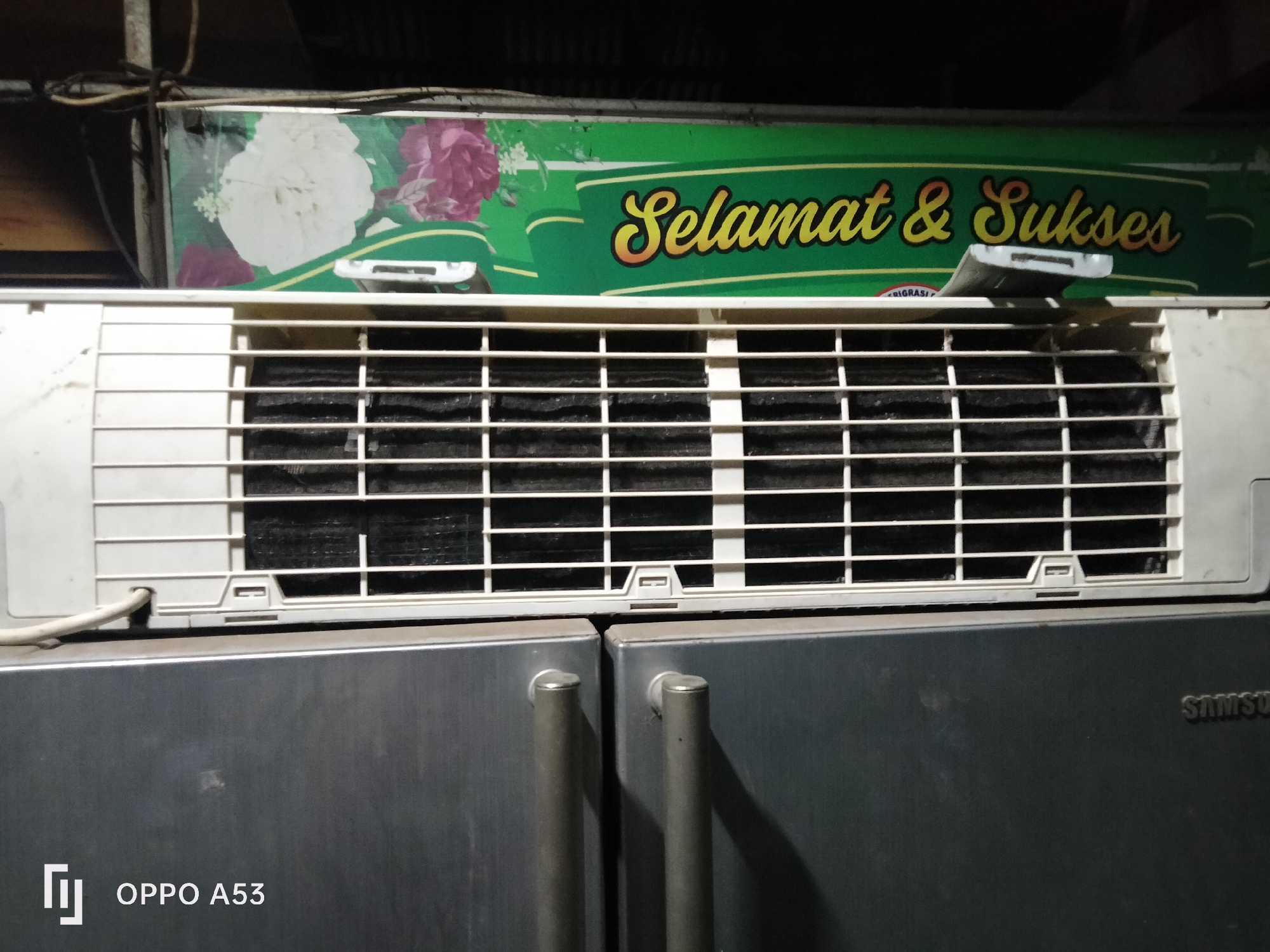 The image shows an air conditioning unit mounted above two refrigerators. The air conditioning unit has a white grille and a green decorative panel with the text "Selamat & Sukses" written in yellow cursive script, alongside images of flowers. The refrigerators appear to be in a kitchen or storage area, with the OPPO A53 logo visible on the left refrigerator door and the Samsung logo on the right. The overall environment appears to be indoors. The image does not provide specific details about the city of Denpasar, Indonesia, beyond the presence of the device and brand names which are common globally. There are no people or interactions depicted.