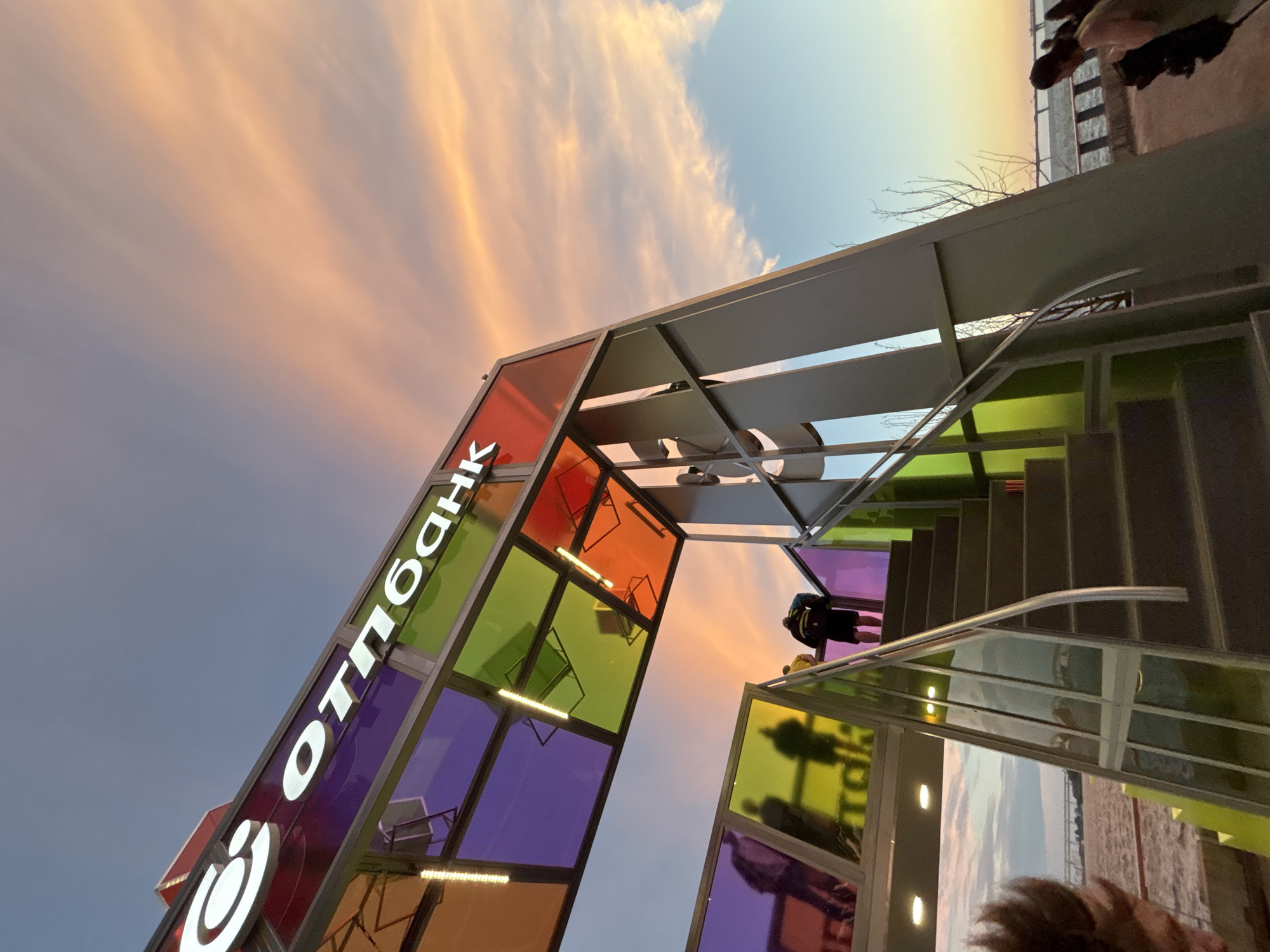 The image captures an outdoor scene at dusk, likely in Saint Petersburg, Russia, given the architectural style and the presence of a body of water. In the foreground, a modern, multi-colored structure with stairs leads upwards. The structure features stained glass panels in shades of purple, green, and orange, illuminated by lights. The logo for "Отпбанк" (OTP Bank) is prominently displayed in white, illuminated lettering on the upper part of the structure.
A person wearing a backpack is ascending the stairs, their back to the camera. Silhouettes of people are visible through the lower stained glass panels. The sky is a mix of blue and a soft, pastel orange, indicating the setting sun, with wispy clouds streaking across it. Beyond the structure, a body of water, possibly a river or bay, is visible, with a bridge or structure in the distance. Other individuals are present in the background, suggesting a public area. The overall atmosphere is serene, with the colorful architecture contrasting with the natural twilight sky.