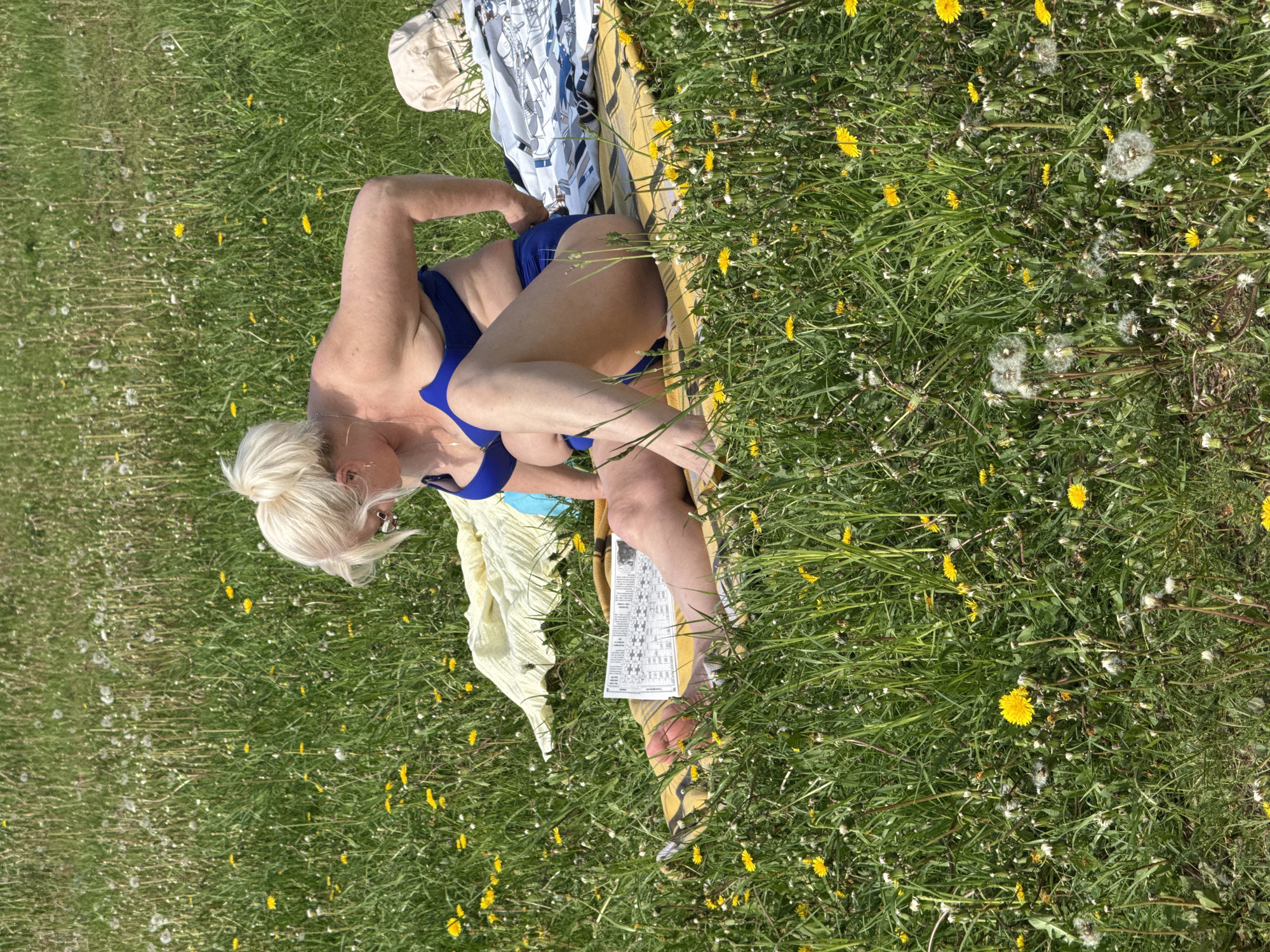A mature Caucasian woman is sitting on a yellow striped towel in a grassy field dotted with dandelions. She is wearing a blue bikini and has blonde hair styled in a bun. Her head is turned toward the left. She is holding a piece of paper. Her belongings, including clothes and a beige hat, are scattered around her. This photo was taken outside in Novosinkovo, Russia.