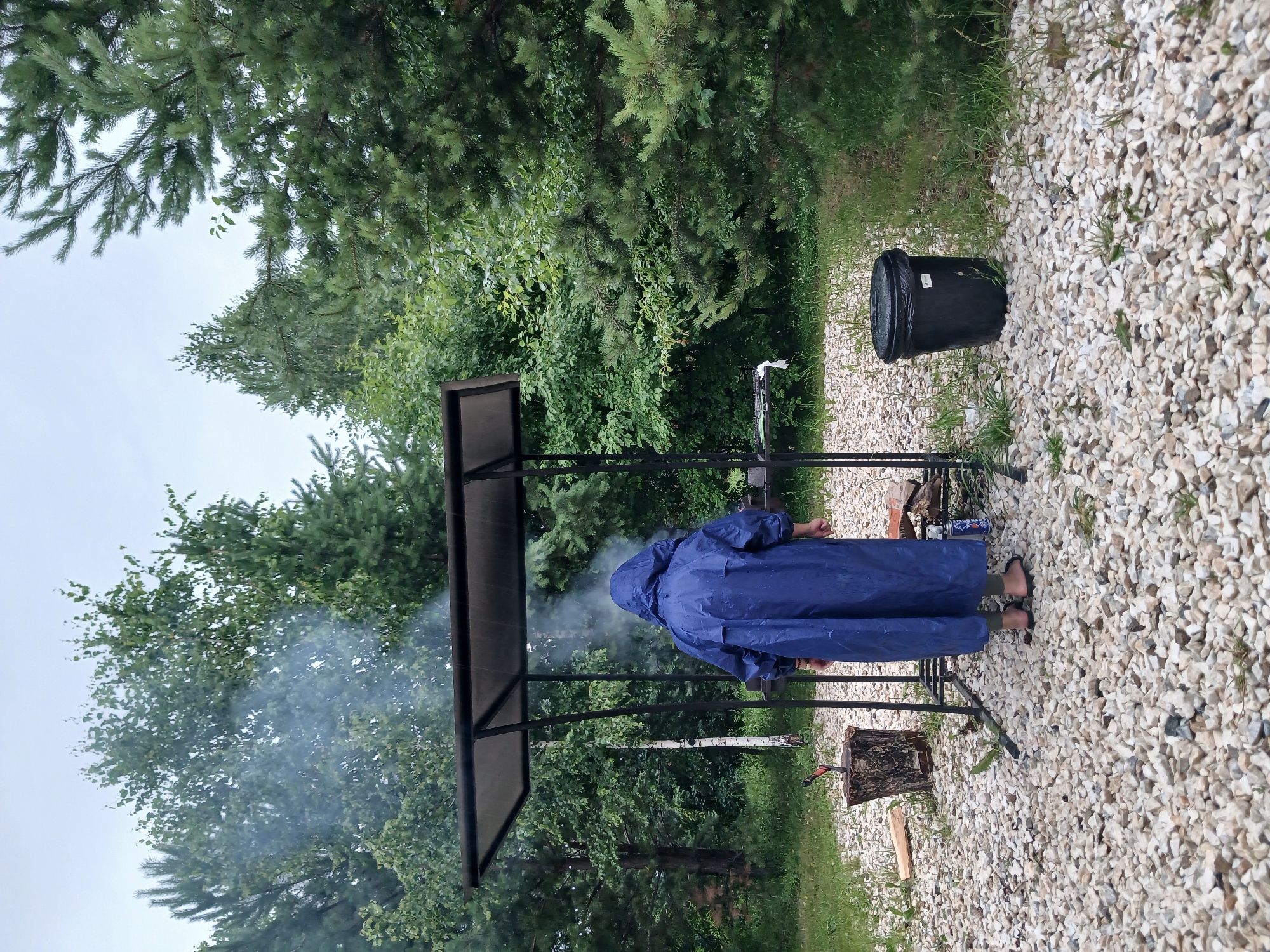 In Verkhneye Dubrovo, Russia, a person wearing a blue hooded raincoat stands in front of a metal grill under a canopy. Smoke emanates from the grill, suggesting food is being cooked. The ground is covered in light-colored gravel, with patches of green grass and small plants interspersed. To the left of the grill, a tree stump with an axe embedded in it is visible, along with a pile of wood. To the right, a black trash can stands on the gravel. The background is filled with dense green trees and foliage, and the sky is overcast. The overall scene depicts an outdoor cooking activity in a somewhat humid or rainy environment, possibly a backyard or a rural setting.