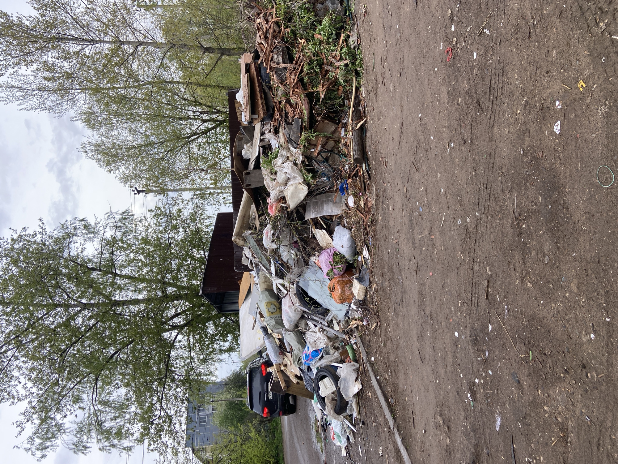 The image shows a large pile of refuse accumulated against a metal structure, possibly a dumpster or shed. The debris includes wood scraps, plastic bags, discarded furniture parts (possibly a mattress), tires, and general household trash. Some vegetation and branches are mixed in with the garbage. The location appears to be an informal dumping site alongside a dirt road or path, with trees and a multi-story building visible in the background. The visible license plate indicates the location of the car to be Russia. From the license plate number, the specific location is determined to be Chaikovsky. The ground is muddy, littered with smaller debris. The sky is overcast. A car is partially visible behind the trash pile.