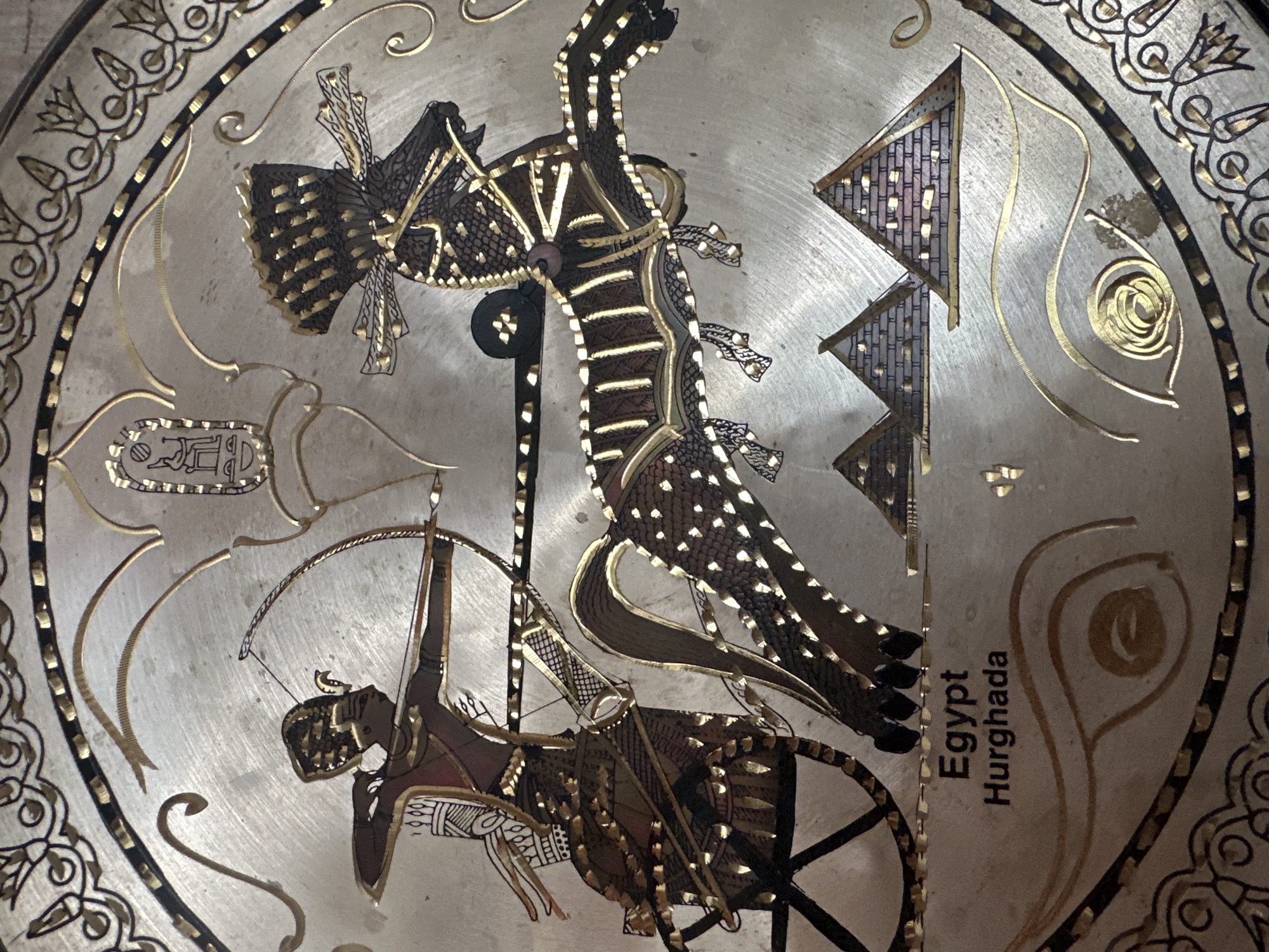 This is a close-up, top-down shot of a decorative plate or platter. The plate is adorned with intricate designs in gold and dark brown, inlaid into a silver-toned metal. The central imagery depicts an Egyptian-themed scene. A figure, possibly an archer, is shown in the lower left corner, drawing a bow. The archer is seated within a chariot or cart with spoked wheels.
Dominating the upper portion of the plate is a large, stylized creature resembling a dragon or mythical bird with wings spread. This creature is rendered with detailed scales and feathers. In the background, abstract triangular shapes are present, also decorated with inlaid patterns. Hieroglyphic-like symbols are visible in the upper left quadrant. Along the right edge of the plate, the words "Egypt" and "Hurghada" are inscribed vertically. The overall aesthetic suggests a blend of ancient Egyptian motifs and possibly some decorative elements influenced by Islamic art, given the swirling patterns and geometric designs. The lighting is even, and there are no indications of weather or time of day, as the scene is on a static object. The location context provided (Пярэжырскі сельскі Савет, Belarus) refers to the administrative region where the image was taken, not the depicted subject matter.