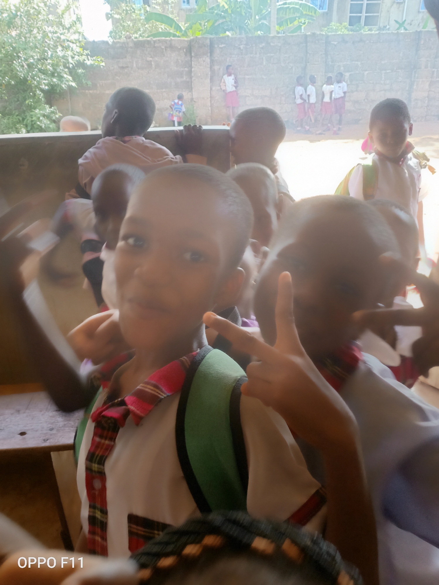 A group of elementary-aged children in school uniforms are gathered. The setting is Ukwu-Abwa, Nigeria, indicated by the photographic device watermark. Children wear uniforms of white shirts with plaid accents and green backpacks. Some children are in the foreground, closer to the camera, posing with hand gestures. Several more are in the background near a concrete wall. Behind the wall are trees and buildings.