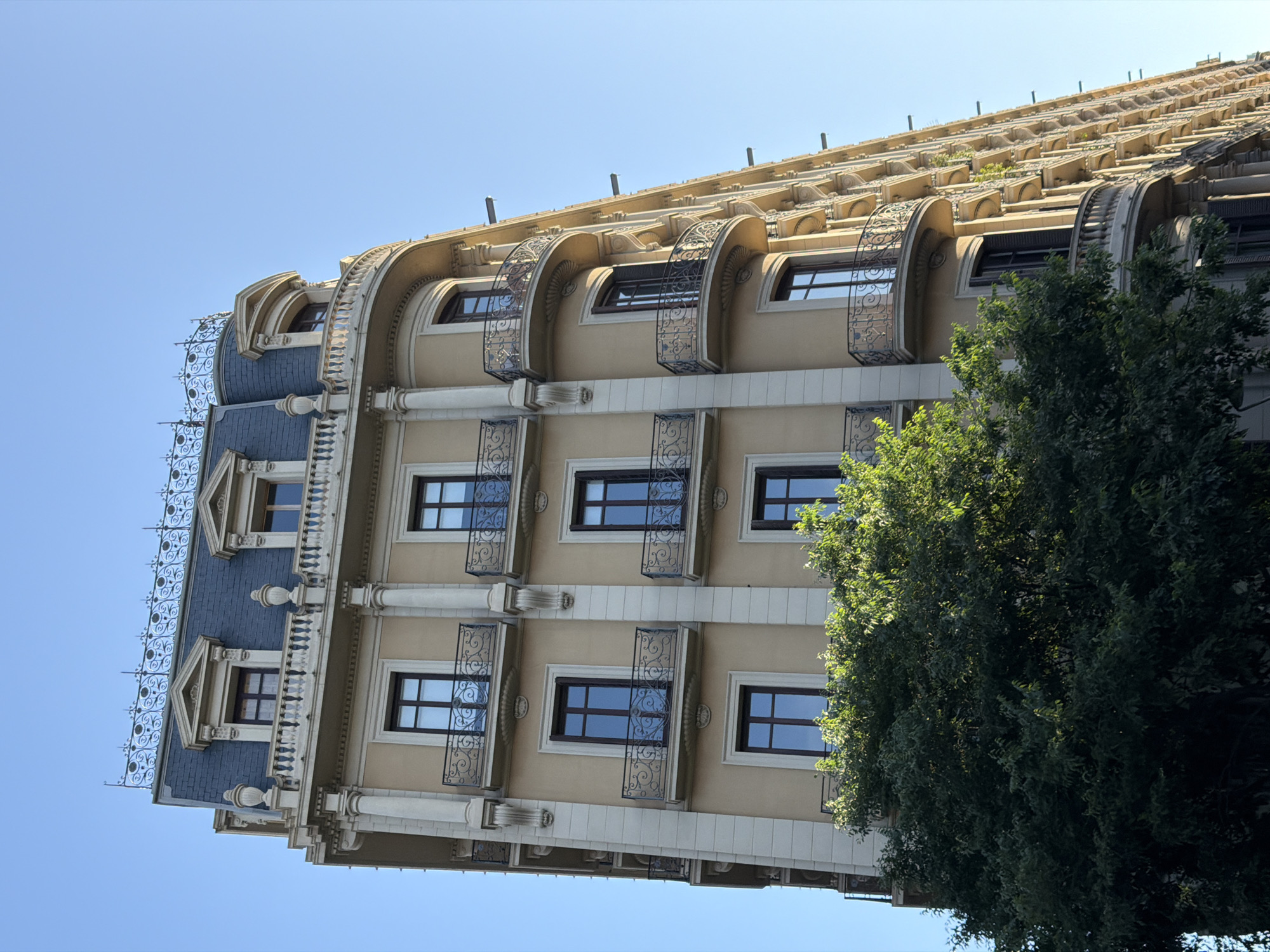 An ornate, multi-story building facade in Barcelona, Spain, is captured under a clear, bright blue sky. The architecture features light beige walls with white trim, dark-framed windows, and intricate detailing. A large, dense green tree partially obscures the right side of the building.
The upper sections of the building display a mansard roof with dark blue/grey tiles and elaborate wrought iron decorative elements along its ridge. Below this, dormer windows are visible, along with multiple levels of rectangular windows, many adorned with decorative wrought iron balconies or railings. The topmost visible floors feature distinct arch-shaped windows, each with a curved, ornate wrought iron balcony. Above these, a terraced or stepped roof design incorporates repetitive architectural elements and some integrated greenery.
The windows reflect the bright daylight, suggesting a sunny day. No people are visible, and there is no discernible text within the image. The image is oriented diagonally, presenting the building at an angle.