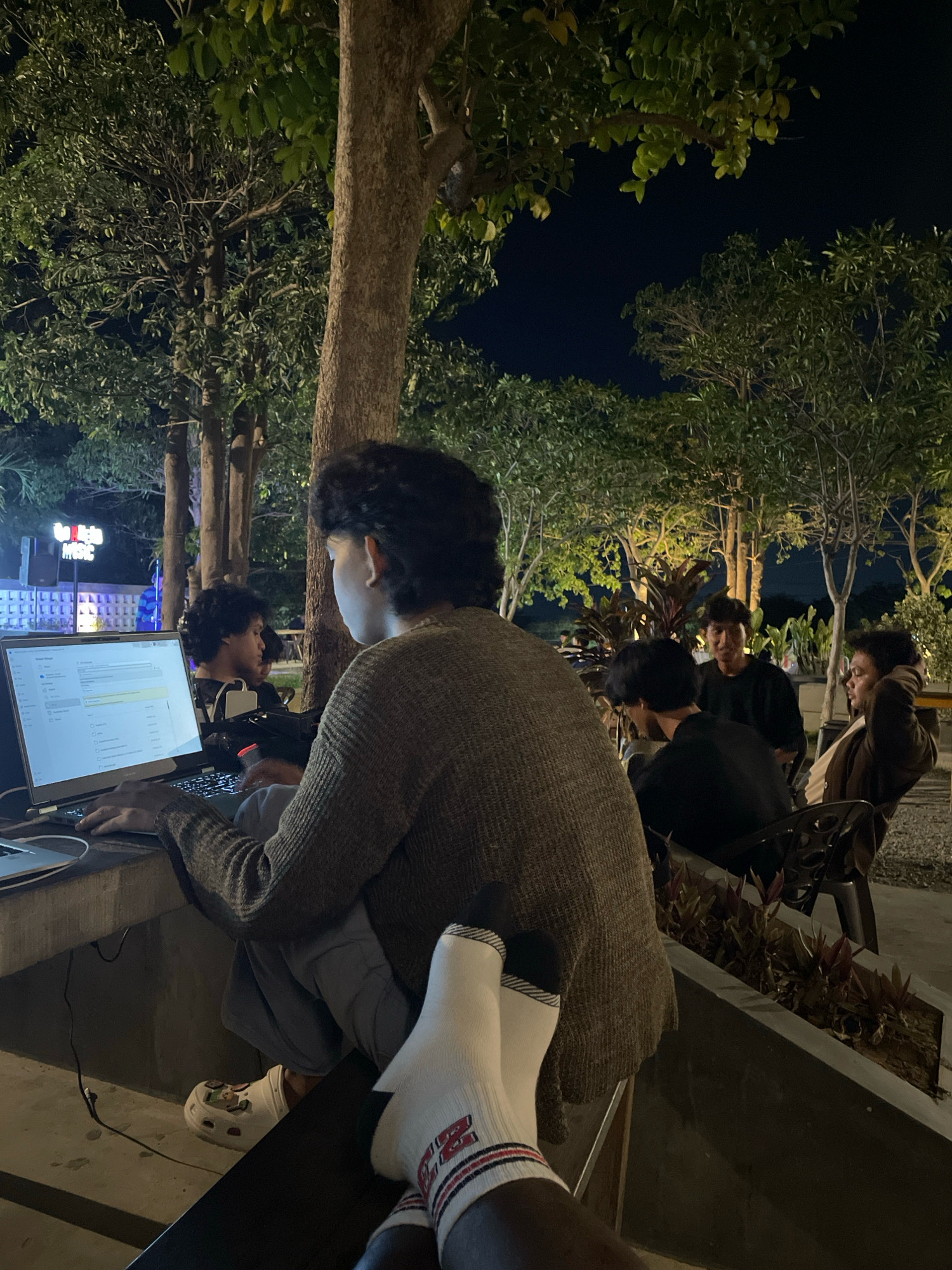 The image depicts a group of young men in an outdoor, dimly lit setting at night. The primary subject is a young man in the foreground, facing away from the camera and towards a laptop. He is wearing a brown knitted sweater, grey pants, and white socks with red and black stripes. His feet are resting on a surface in front of him, showing off his colorful socks. The laptop is open and illuminated, suggesting he is working or engaged with it.
Behind him, a few other young men are visible. One is sitting and looking towards the laptop, another is seated and looking slightly to the side, and a third is leaning back in a chair with his arm behind his head, looking towards the camera. They are all dressed casually.
The environment appears to be a courtyard or an outdoor seating area, possibly a cafe or restaurant. There are trees with foliage casting shadows and providing a natural backdrop. In the background, there's a glimpse of a brightly lit structure with signage that reads "MUSIC". The lighting in the scene is a mix of ambient streetlights, the glow from the laptop, and possibly some decorative lighting. The overall impression is a casual, late-night gathering with a focus on technology and social interaction. The context of Talise, Indonesia, suggests a tropical or subtropical climate, which might explain the outdoor setting.
