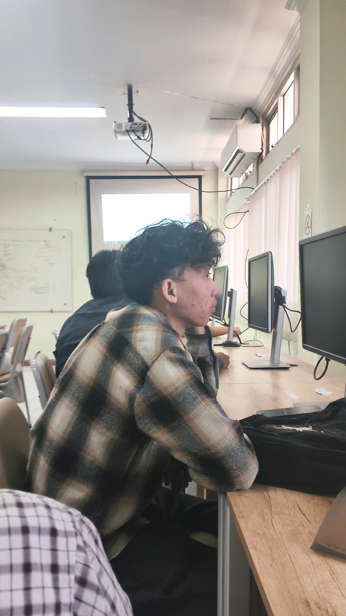 The image depicts an indoor scene, likely a classroom or computer lab, with several young men seated at desks with computer monitors. The primary subject is a young man in the foreground, viewed in profile, wearing a plaid shirt. He has curly dark hair and appears to be looking at a computer screen. Other individuals are visible in the background, also seated at desks with computers. The environment includes desks, monitors, chairs, and a projector mounted on the ceiling, suggesting an educational setting. The lighting appears to be artificial, coming from overhead lights and possibly natural light from the windows. The overall atmosphere seems to be one of study or work. The specific location of Lohbener, Indonesia, cannot be determined from the visual information present in the image.