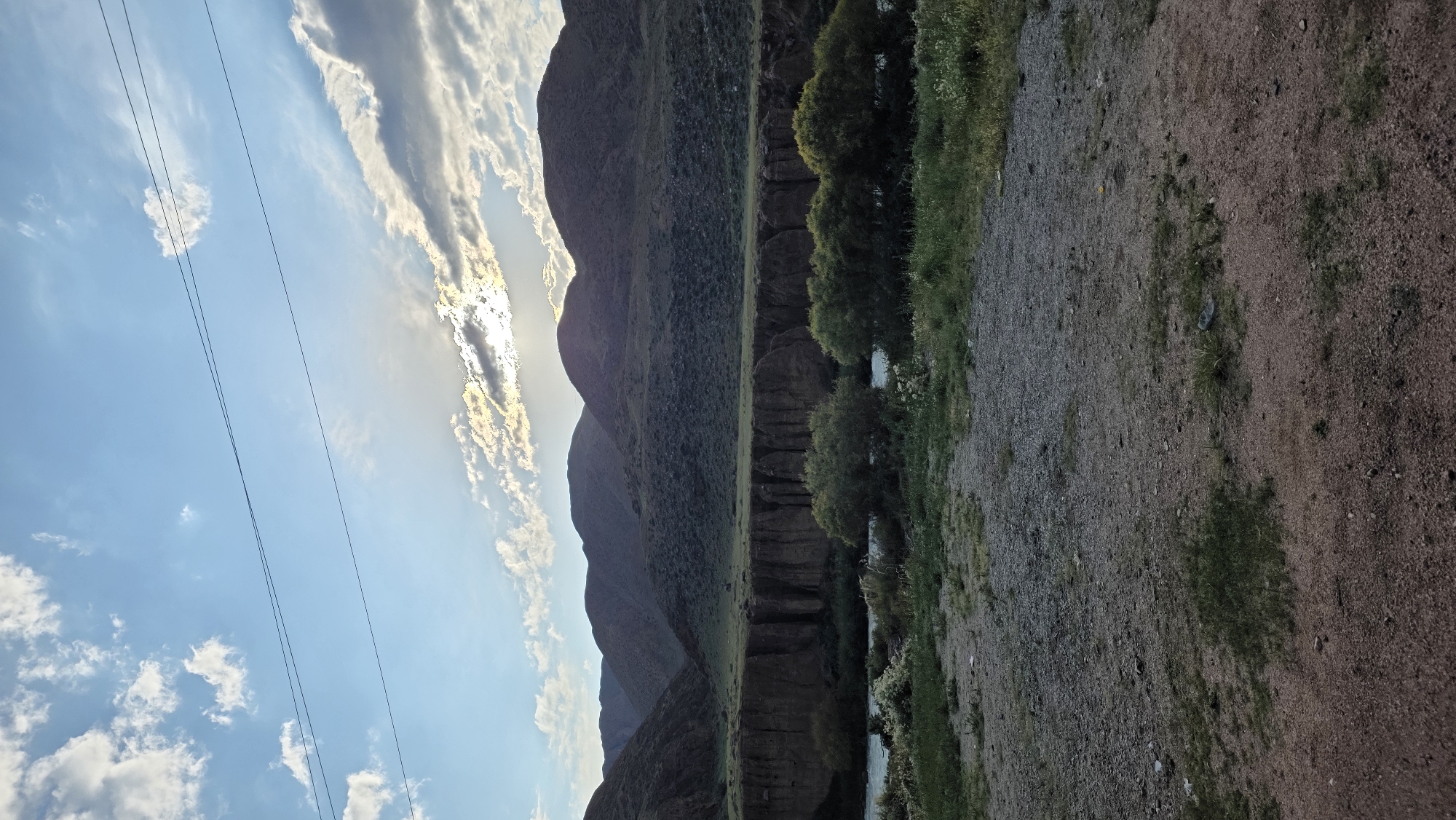 The image depicts a natural landscape featuring a river flowing through a canyon with eroded cliffs. The canyon walls are a reddish-brown color, exhibiting vertical striations. Above the canyon, rolling, green hills rise up to meet a sky with scattered white and gray clouds. The sun is partially obscured by clouds, casting a bright glow in the upper right portion of the sky. Two thin, dark power lines stretch horizontally across the sky in the upper section of the image. In the foreground, the ground is covered with gravel and sparse green vegetation. The scene appears to be located in a rural or mountainous region. There are no people or animals visible in the image.