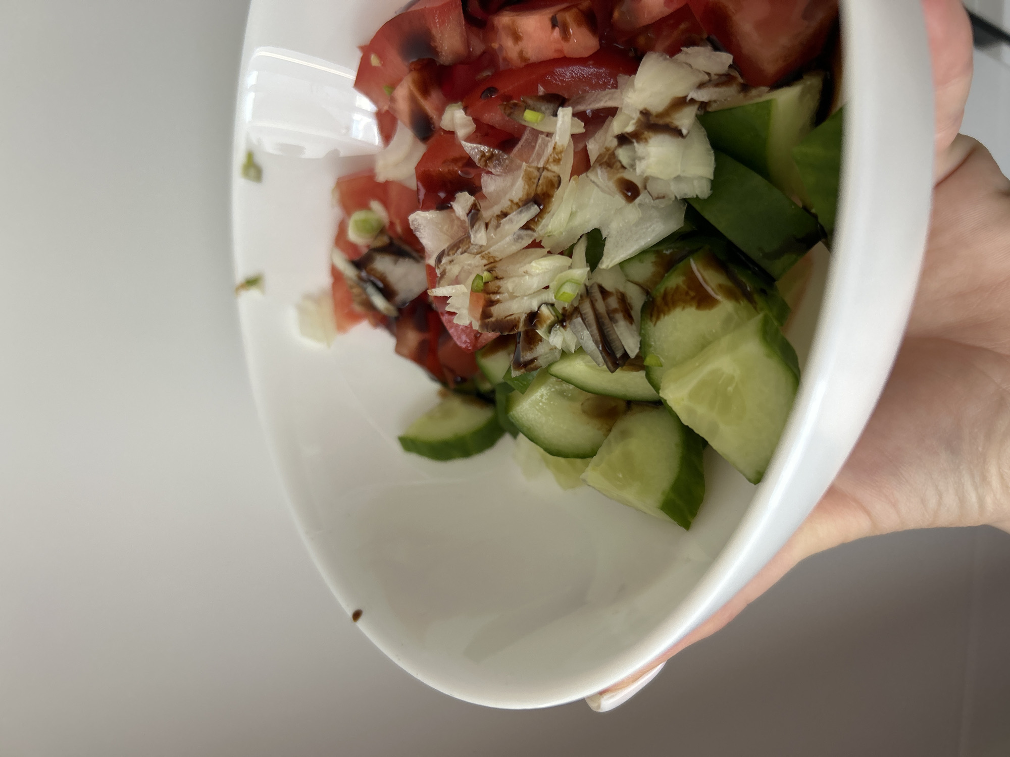 A white bowl held by a human hand contains a salad composed of diced tomatoes, sliced cucumbers, shaved white cheese, and chopped green onions, dressed with a dark brown sauce, likely balsamic glaze. The scene is indoors. There is no evidence to indicate the location is in Chaikovsky, Russia.