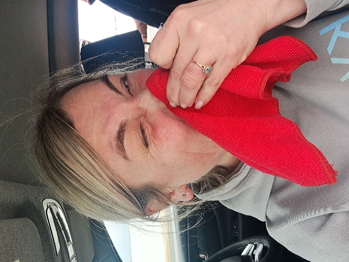 An adult female, with light-colored hair and visible facial redness around the nose and cheek, is depicted inside a vehicle, specifically in the city of Sasovo, Russia. She is wearing a light gray hooded sweatshirt and holds a folded red microfiber cloth against her nose and mouth area with her right hand. Her right eye is partially visible and squinted, while her left eye is closed, indicating a strong sensory reaction, possibly a sneeze or irritation. A gold-toned ring with a light-colored gemstone is visible on her right ring finger. The vehicle's interior features a gray headliner, an overhead light fixture, and part of a black steering wheel on the left. A dark-clad figure or object is partially visible through a window in the right background.