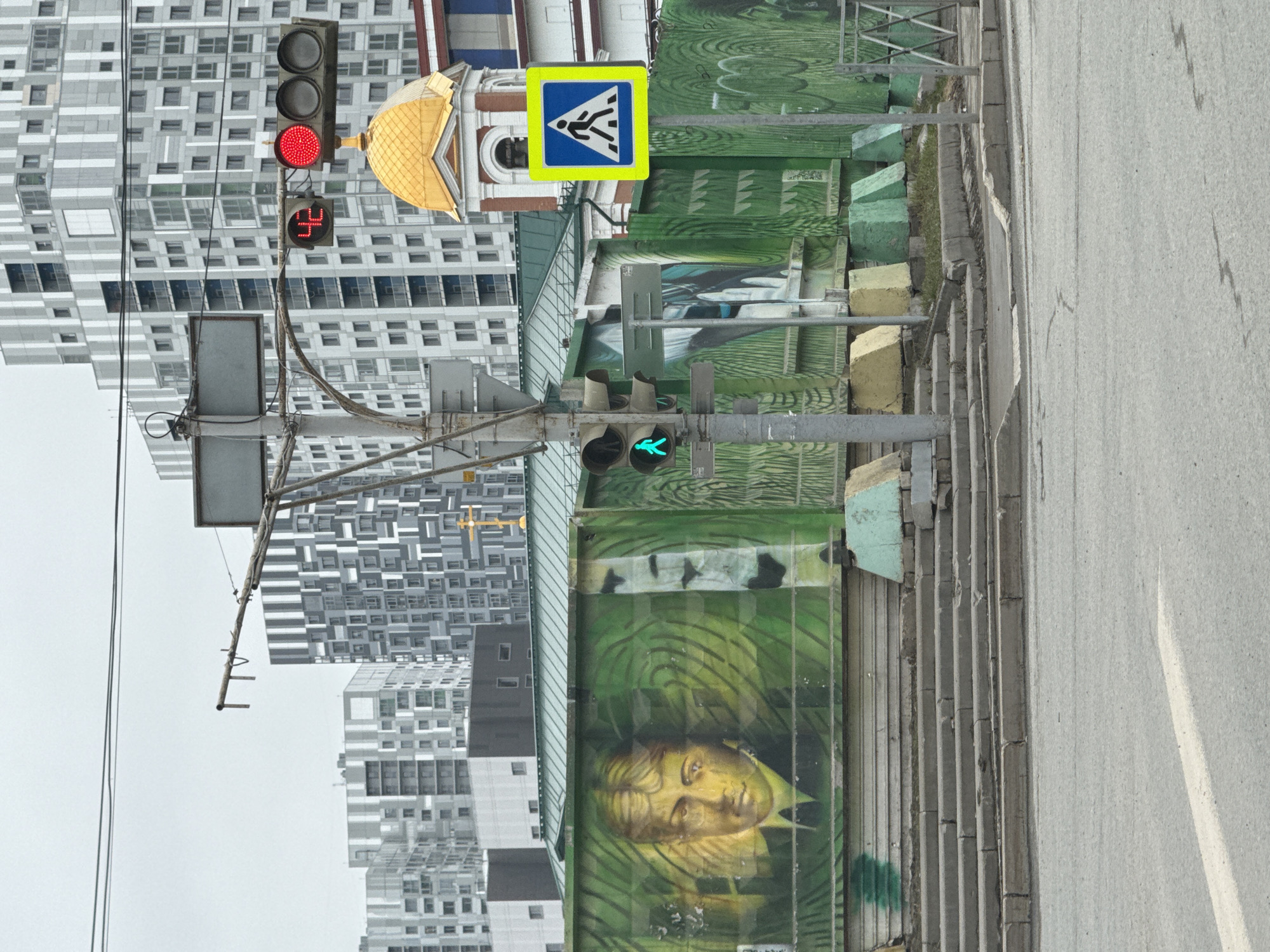 The image shows a street scene in Perm, Russia. A pedestrian crossing sign is visible above a fenced area decorated with a mural of a face. A traffic light shows a green walking figure, indicating it's safe for pedestrians to cross. Above that traffic light is a traffic light for vehicles that is red, along with a numerical display. The scene is framed by modern apartment buildings in the background and the edge of a street to the right.