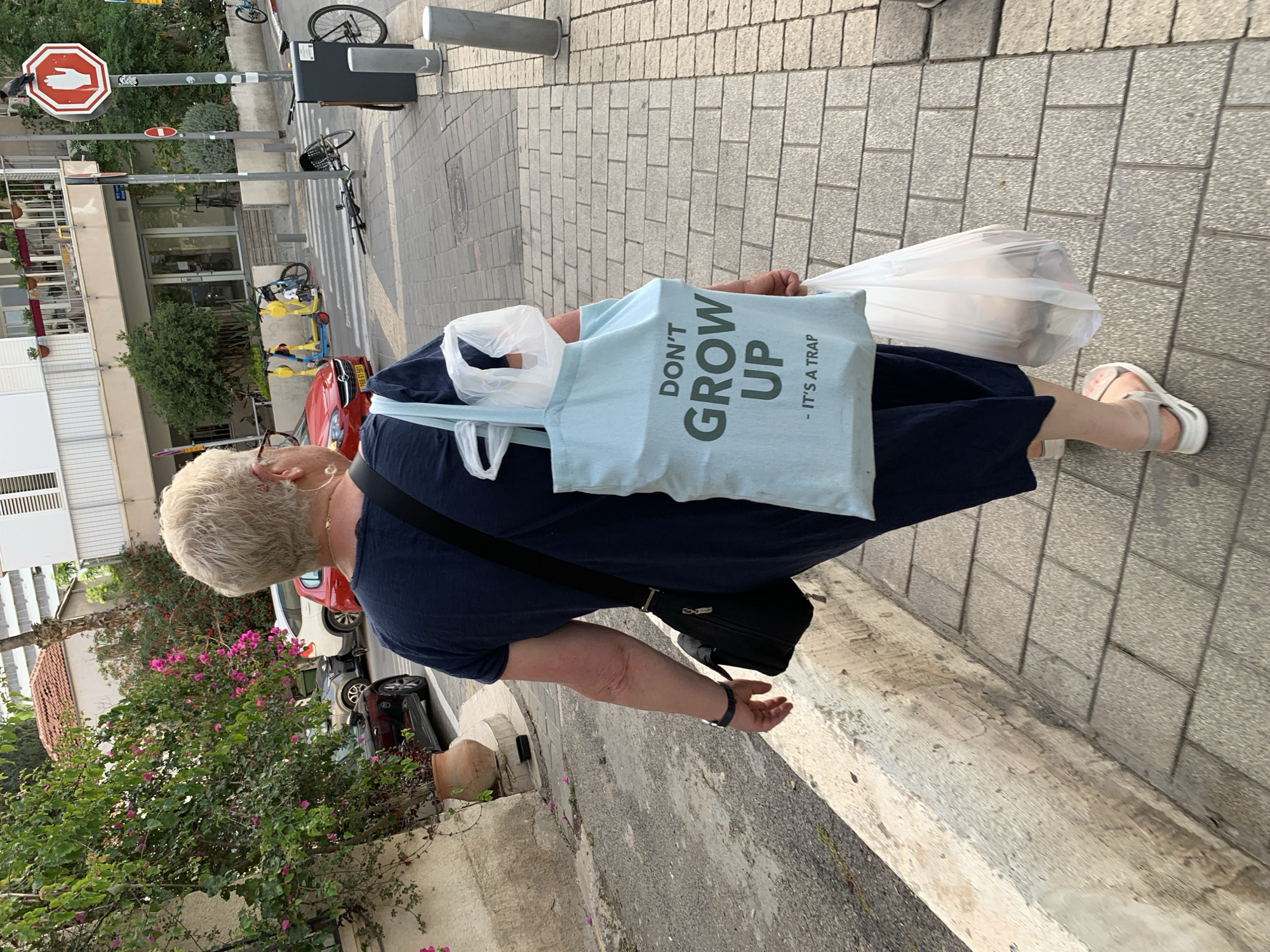 A woman is walking down a brick sidewalk in Tel-Aviv, Israel. She has short blonde hair and wears a dark blue dress and grey sandals. She is carrying a light blue tote bag with the words "DON'T GROW UP - IT'S A TRAP" printed on it, as well as two white plastic bags. She is wearing a black cross-body bag and a watch. A stop sign is visible in the background. A red car is parked on the side of the road. A bougainvillea plant with pink flowers grows nearby.