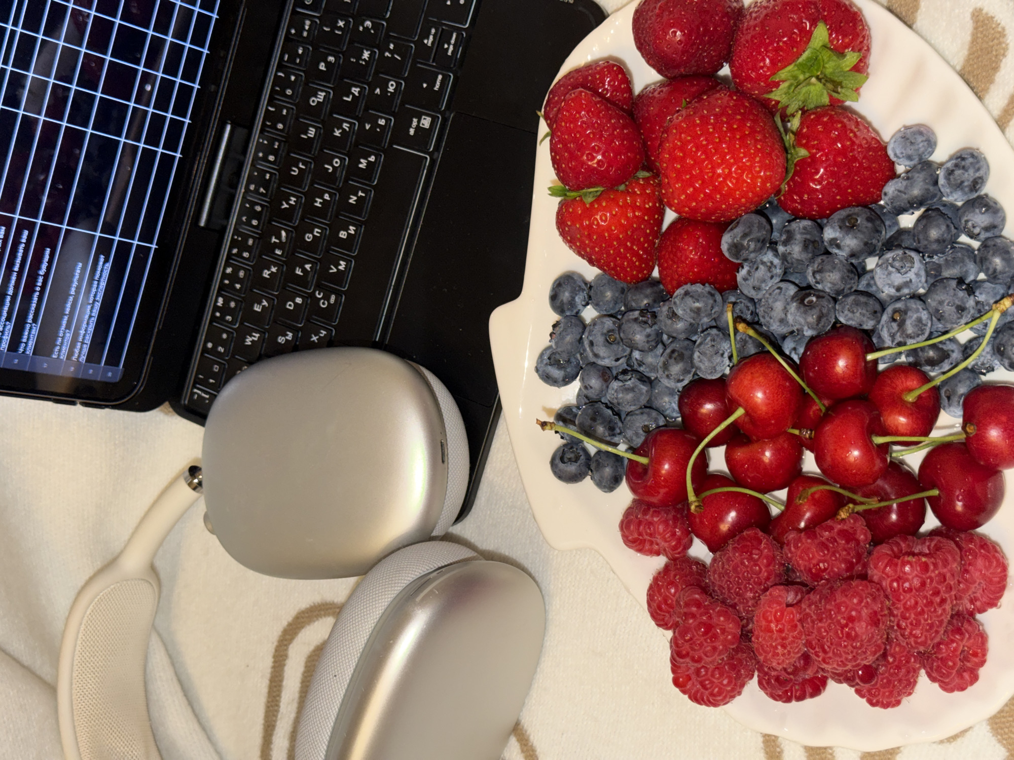 The image shows a close-up of a plate filled with fresh berries: raspberries, blueberries, cherries, and strawberries. Behind the plate, a pair of silver headphones rests on a textured, off-white surface. To the right of the headphones, a black laptop is partially visible, with its keyboard illuminated and a spreadsheet displayed on the screen. The scene is brightly lit, highlighting the vibrant colors of the fruit and the metallic sheen of the headphones. The background appears to be a soft, neutral-colored fabric. There is no indication of a specific location in Moscow, Russia, or any people present in the image.