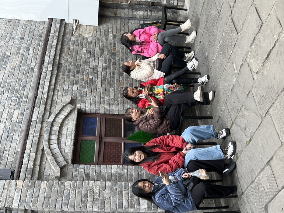 Six adult females are seated in a row on black metal chairs against a gray brick wall in Yuzhong District, China. The wall features an arched window with a brown frame, containing four panes of colored glass: green and blue on top, and two patterned brown/red panes below. The ground is paved with uneven gray stone slabs.
From left to right:
1. A female with long dark hair, wearing a blue quilted jacket and black pants, holds a white smartphone in her right hand, looking at the screen. Her left hand holds a transparent plastic bag containing light-colored food items. A black shoulder bag is visible on her lap.
2. A female with long dark hair, wearing a red puffer jacket and light blue jeans, sits with her hands resting in her lap, looking forward.
3. A female with short dark hair, wearing a brown patterned quilted jacket and black pants, holds a yellow-capped bottle in her right hand and looks to her right, away from the camera.
4. A female with dark hair pulled back, wearing sunglasses on her head, a red jacket, and a multi-colored scarf, holds a white smartphone with both hands, looking at the screen. She wears black leggings.
5. A female with dark hair pulled back, wearing a beige puffer jacket and black pants, sits with her hands resting in her lap, looking to her right, away from the camera.
6. A female with long dark hair, wearing a bright pink puffer jacket and gray leggings, sits with her hands clasped in her lap, looking to her right, away from the camera. She wears white ankle boots.