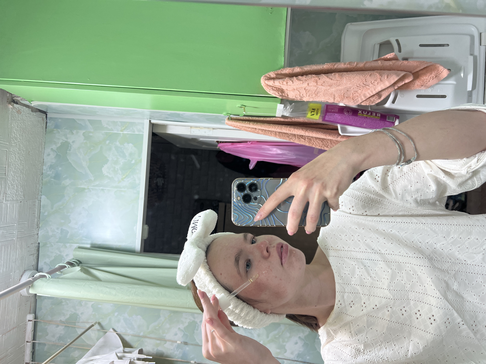 A person is taking a selfie in a bathroom. The person is wearing a white top and a white headband with bunny ears. They are holding a dropper with a clear liquid near their face. The bathroom has light green cabinets, patterned wallpaper, and a drying rack with clothes. A pink towel and a package labeled "RELUX" are visible on a shelf. The location is Chaikovsky, Russia.