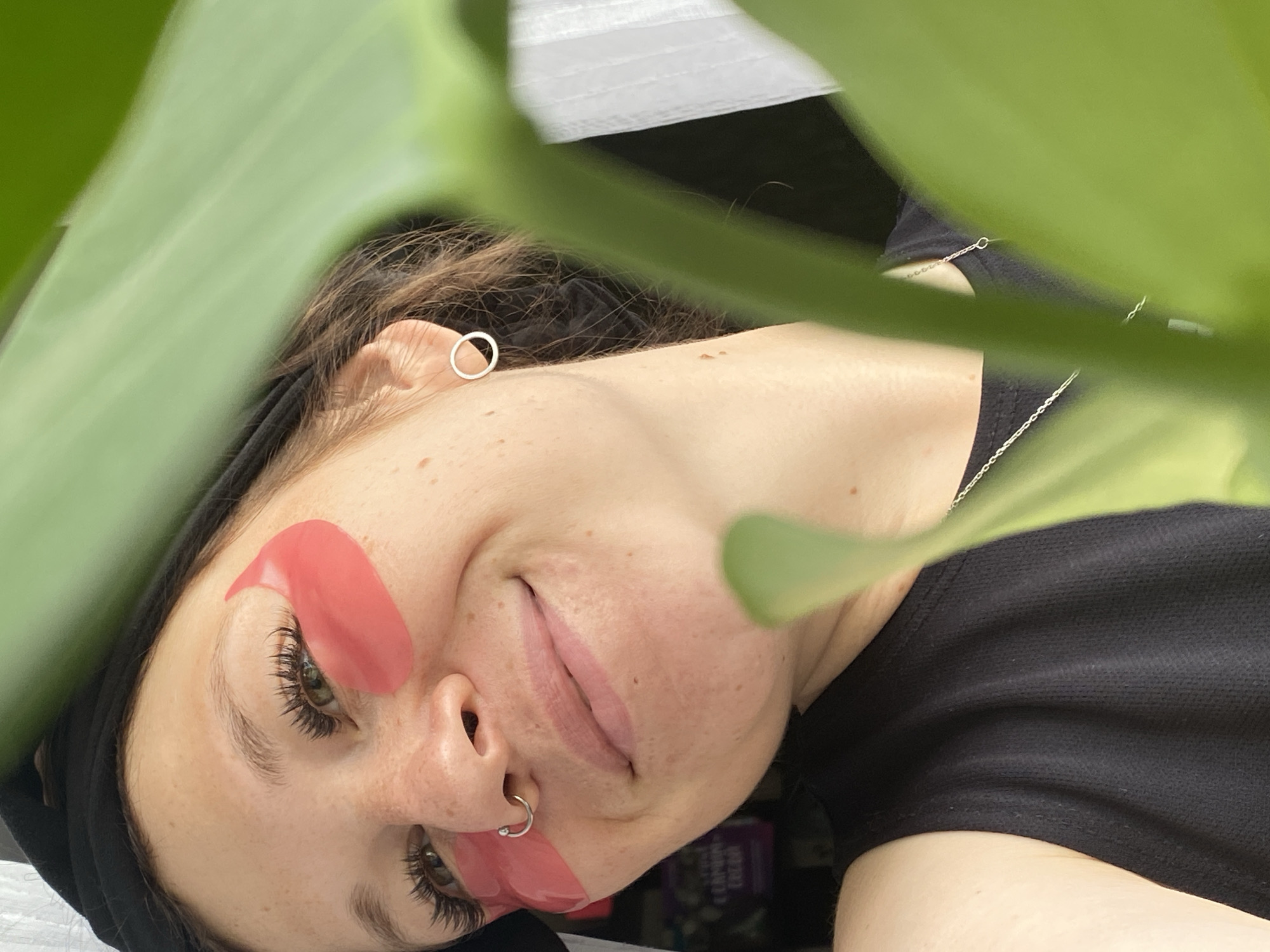 The image shows a close-up of a young woman wearing pink under-eye patches and a black headband. She has long eyelashes, a nose ring, and a small silver hoop earring. She is smiling and looking to her left. A large green leaf is in the foreground, partially obscuring her face. The background is blurred, but appears to be an indoor setting with some shelves visible. There is no information in the image to suggest it is located in Chaikovsky, Russia.