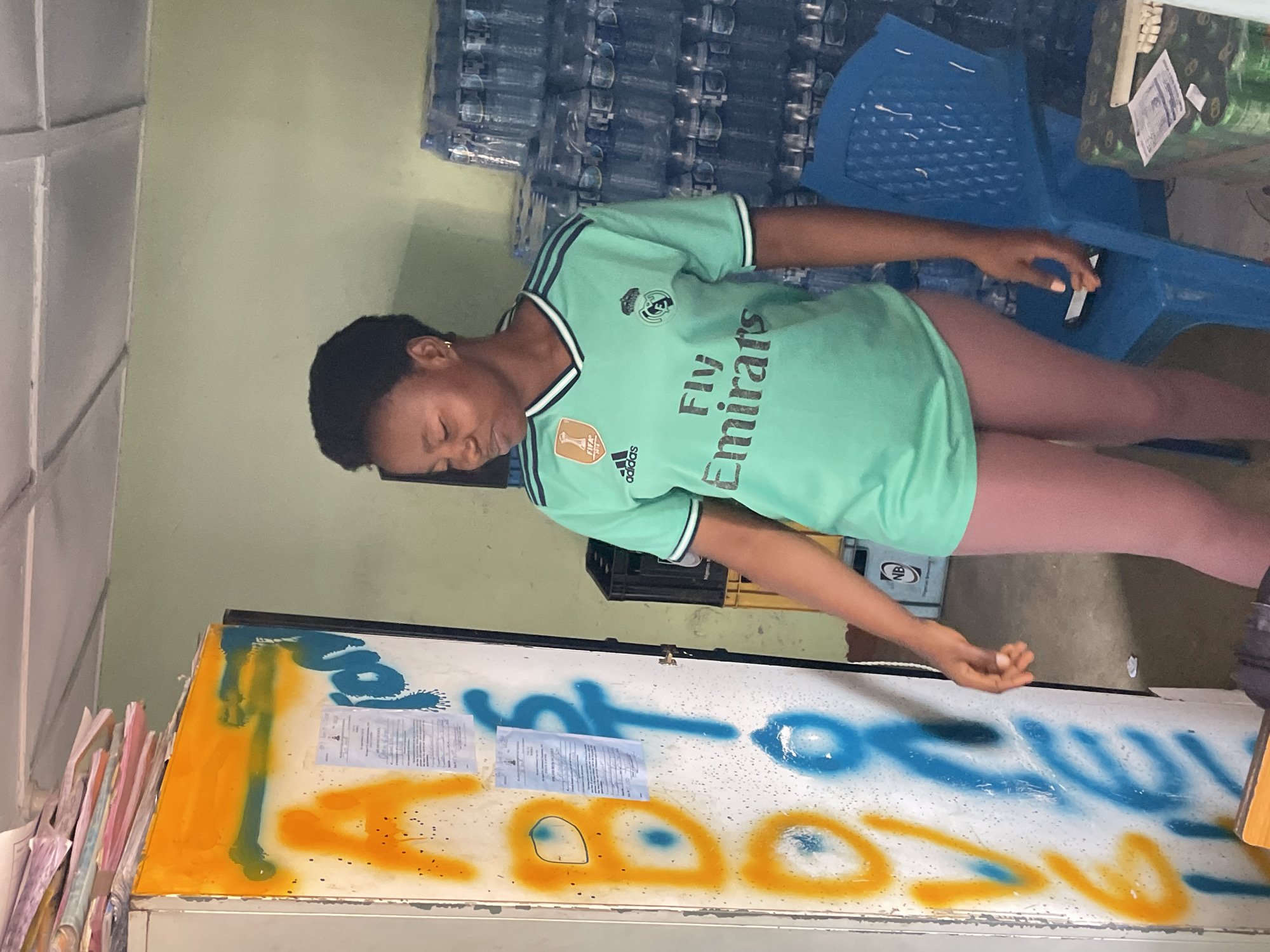 The image depicts an indoor scene, possibly a shop or storeroom, in Jos, Nigeria. A young woman wearing a light green Real Madrid jersey and pink leggings stands in front of a white metal cabinet that is covered in colorful graffiti. The graffiti consists of large, bold letters in orange and blue, as well as some papers attached to the cabinet door. To the right of the woman, stacks of bottled water are visible, along with a blue plastic chair. A black crate and some yellow crates are also present in the background. The overall impression is that of a functional, somewhat cluttered commercial space.