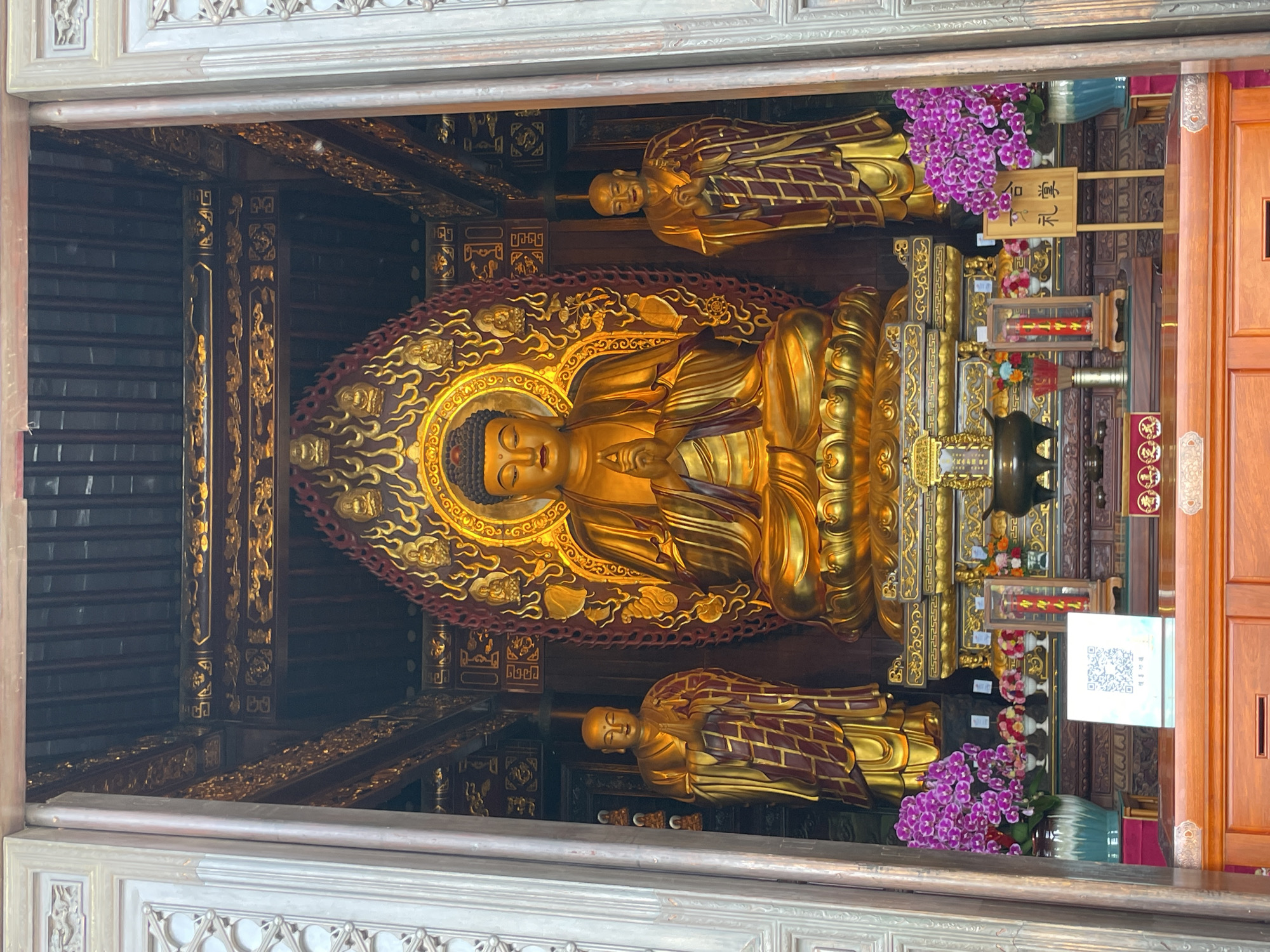 The image depicts a golden Buddha statue flanked by two smaller, gold-colored figures in what appears to be a temple, as evidenced by the intricate woodwork and ornamentation. The setting is within Yanta District, China, as the image recognition system has associated this temple with that location. The Buddha statue is the focal point, surrounded by a golden halo and seated on an ornate platform. Purple flowers adorn the space near the two smaller figures. Behind the statues, there are various religious objects and offerings. The view of the scene is framed by the temple's doorway, which exhibits detailed carvings.