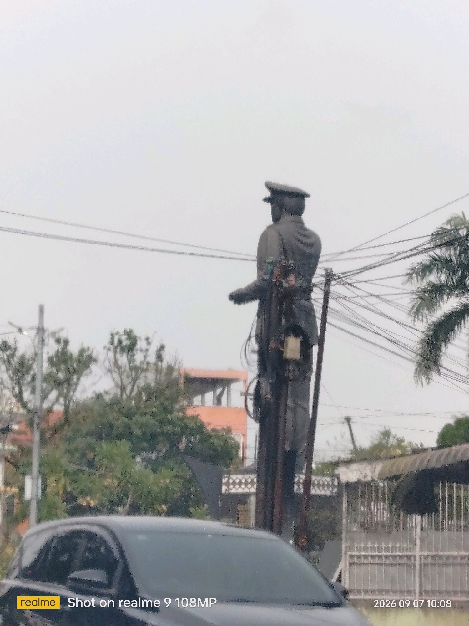 The image shows a statue of a person in uniform, possibly a military or police officer, standing in an urban environment. The statue is dark in color and appears to be made of metal. It is positioned in front of a background of trees, buildings, and power lines. In the foreground, a portion of a dark-colored car is visible. The sky is overcast and appears to be a light gray color. The scene suggests a public space, potentially a street or a park, within the City of Medan, Indonesia, given the timestamp indicating the date and time. The statue is the central focus, with its gaze directed to the right of the frame. There are electrical wires and poles surrounding the statue, indicating an active, developed area. The overall impression is one of a slightly hazy or cloudy day in a bustling city.