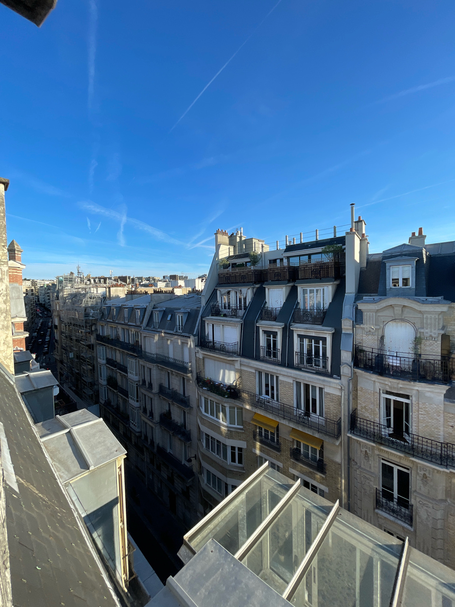 This is an elevated, daytime, eye-level view of a street in Paris, France, captured on a clear, sunny day with a bright blue sky and wispy white contrails. The scene is dominated by the architecture of Parisian buildings, characterized by their mansard roofs, dormer windows, and intricate facades. The buildings are densely packed along a street that recedes into the distance.
In the foreground, a portion of a rooftop is visible on the left, with a grey, pitched surface and a metallic vent. Adjacent to this is another angled roof section, partially covered by a dark, flat surface, and a lighter grey, box-like structure. Further to the right, a glass-covered structure with a metallic frame occupies a significant portion of the foreground, suggesting a rooftop extension or conservatory.
The midground is comprised of several multi-story apartment buildings. The closest building on the right features a light-colored, textured facade with ornate architectural details around a large window. This building has a black wrought-iron balcony, and a window is ajar, revealing a glimpse of the interior. The building to its left displays a lighter brick facade with multiple windows, many of which are tall and narrow, also featuring black wrought-iron balconies. Yellow awnings are visible over some windows on a lower floor of this building. The buildings continue in a line towards the back, with varying facade colors and architectural styles.
The background shows a continuation of urban architecture, with many buildings of similar height and style. In the distance, construction scaffolding is visible on one building, indicating ongoing renovation or development. The street below is lined with parked cars, suggesting a typical urban streetscape. The overall impression is one of a classic Parisian cityscape, likely in a residential or mixed-use district. There are no people visible in the frame.