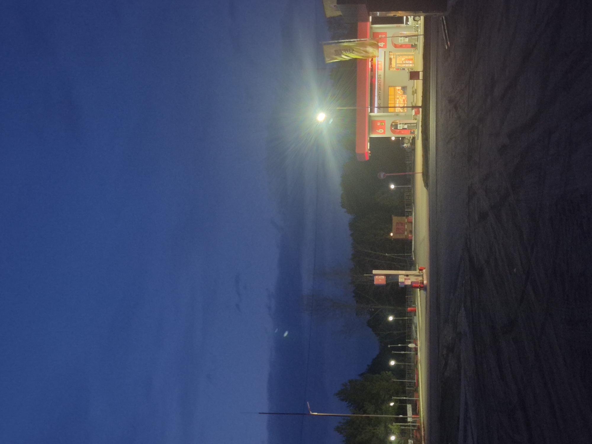 The image, taken in Chaikovsky, Russia, depicts a gas station at night or late dusk. The scene is dominated by a deep blue sky that transitions from lighter shades towards the horizon, with subtle cloud formations. A bright, star-like light source, likely a streetlamp or part of the station's lighting, emits a strong lens flare, illuminating the left side of the frame.
On the right, a brightly lit gas station structure is visible, featuring multiple fuel pumps. The building and pumps are red and white, with yellow interior lighting emanating from its windows. Above the pumps, prices or fuel grades are displayed, with numbers like "92", "95", and "100" clearly visible, indicative of gasoline types. Cyrillic text "АЗС ЭНТУЗИАСТОВ" (AZS ENTUZIASTOV), meaning "Enthusiasts Gas Station," is prominently displayed on the station canopy or building facade. Numbers "4" and "6" are also visible on what appear to be individual pump stations.
In the mid-ground, a line of dark trees or bushes is silhouetted against the sky, separating the gas station area from the vast open sky. A paved dark surface, likely a road or parking lot, occupies the lower right portion of the image, showing some tire marks or patterns. Several smaller streetlights line this paved area. No people or active vehicles are visible, suggesting a quiet moment at the station despite its illuminated appearance, indicating it is open for business. The overall mood is quiet and still, emphasizing the artificial glow against the natural darkness.