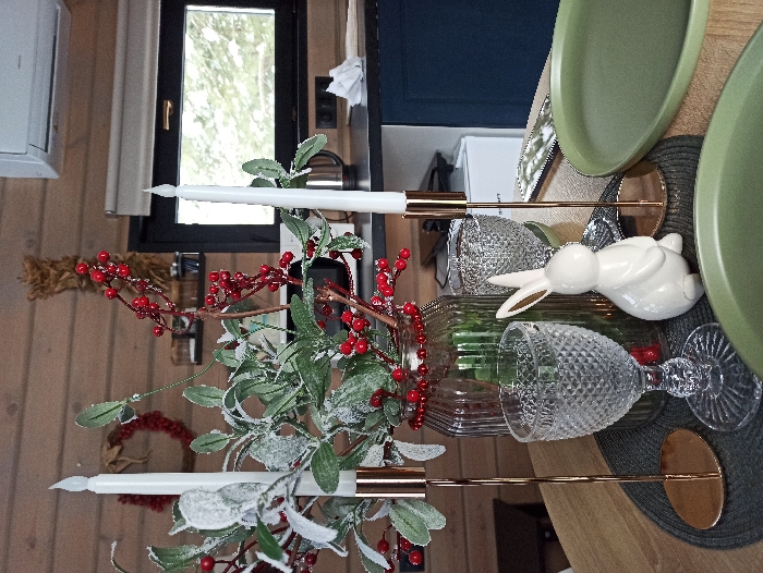 This image depicts a festive table setting inside a residential room in Nizhneye Zadolgoye, Russia. The room features light brown wooden plank walls. In the foreground, a round wooden table is set with a dark grey woven placemat. A clear, ribbed glass vase serves as a centerpiece, containing artificial green foliage, some frosted with white, and clusters of bright red berries. A string of small red beads is wrapped around the vase's neck. Flanking the centerpiece are two slender, white, pointed faux candles (likely LED) in tall, golden-colored metallic candlestick holders. A clear glass goblet with a diamond-patterned texture is positioned to the left of the vase. To the right of the vase, a small, glossy white ceramic rabbit figurine faces left, and another patterned glass is partially visible behind it. Two matte sage-green dinner plates are partially visible in the bottom right and bottom center of the frame.
The background reveals a kitchen or utility area with a dark countertop. On the countertop, a stainless steel electric kettle is visible, along with a crumpled piece of white paper or tissue. A window with a black frame offers a view of an outdoor winter scene with trees. Above the window, an air conditioning unit is mounted on the wooden wall. Additional wall decorations include a red berry wreath to the left and a vertical dried straw arrangement above the centerpiece. No individuals are present in the scene, and no actions or interactions are depicted.