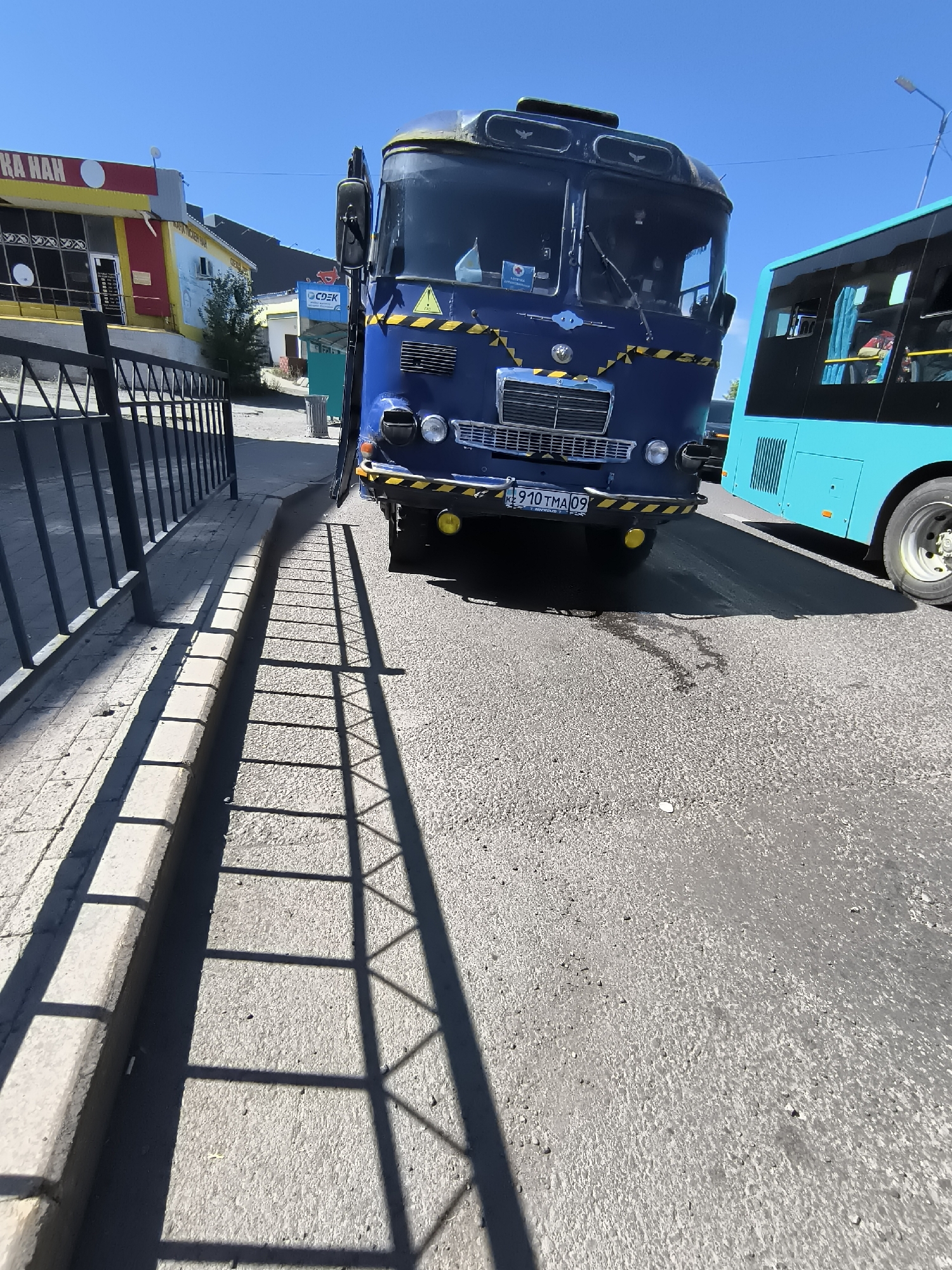 The image shows a street scene in the city of Qarağandı, Kazakhstan. In the foreground, a blue bus is parked on the right side of the road. The bus has yellow and black striped warning tape on its front bumper. Behind it, a turquoise bus is visible, also parked on the road. To the left of the road, there is a sidewalk with a black metal fence and a curb. A building with a yellow and red facade is visible behind the fence. The sky is clear and blue, indicating a sunny day. The pavement is asphalt, and there are some dark stains on the road in front of the blue bus, possibly from a leak. The shadow of the fence is cast across the sidewalk and road.