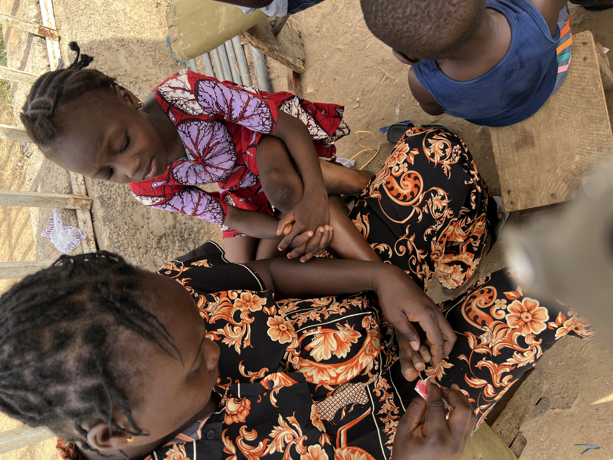 The image shows an outdoor scene in Minna, Nigeria. A child sits on the lap of an adult woman who is seated on the ground. The child wears a red and purple patterned dress. The woman is wearing a black and orange patterned shirt. The woman appears to be examining or treating the child's fingers. Another person is visible in the background wearing a blue tank top and sitting on a wooden bench. The ground is sandy and littered with small debris. A metal gate or fence is partially visible. A tan sack or bag is visible in the upper left corner. The scene appears to be in a residential or public area.