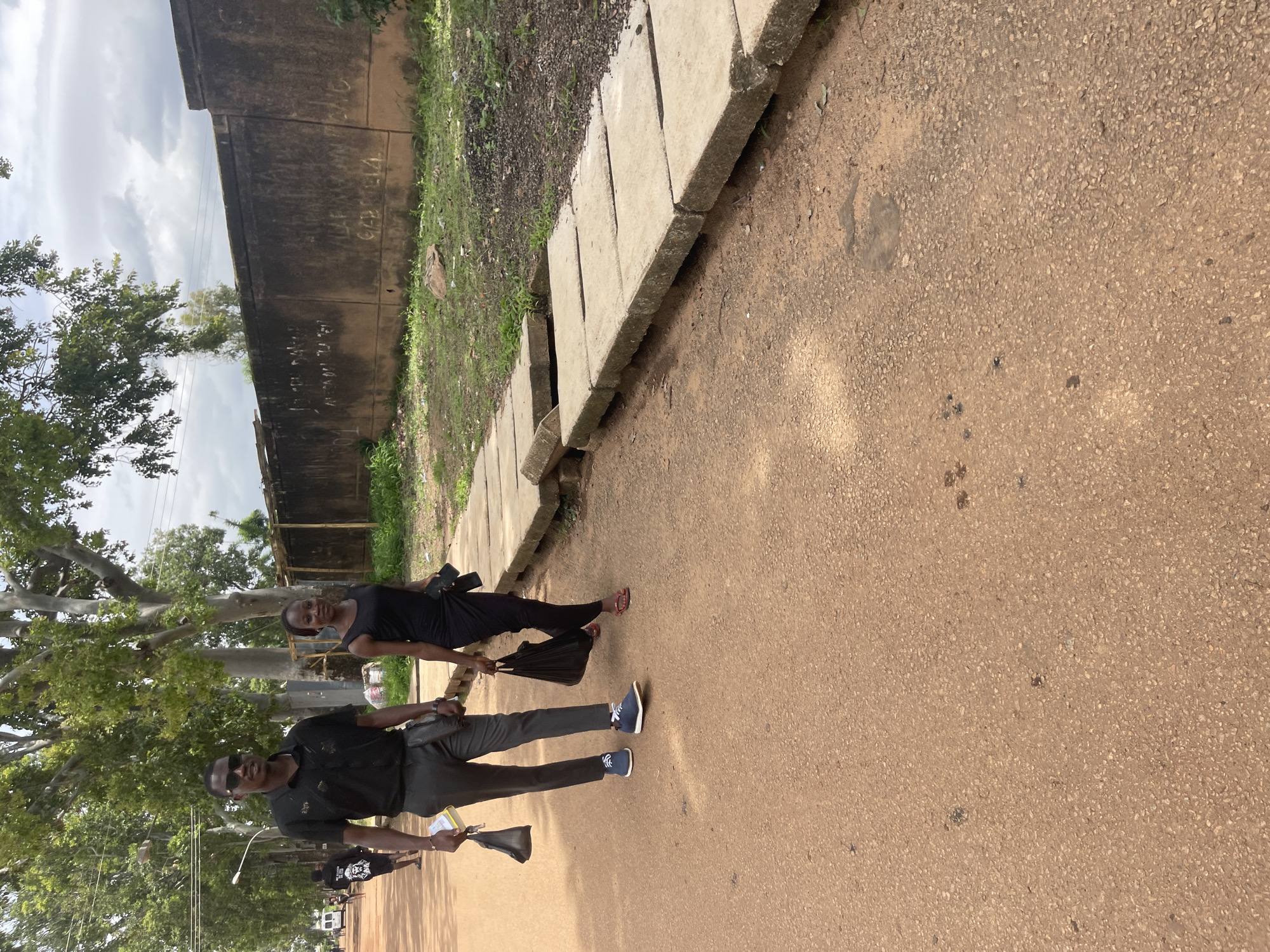 A man and a woman are walking on a dirt road lined with concrete slabs. The man, wearing sunglasses, a black polo shirt, and dark trousers, carries a black bag in his left hand and a booklet in his right. The woman, in a black sleeveless dress, carries a black bag in her left hand and a wallet in her right. In the background, a concrete wall is visible with graffiti. Trees and greenery surround the scene. The image was taken in Rafin Par, Nigeria.
