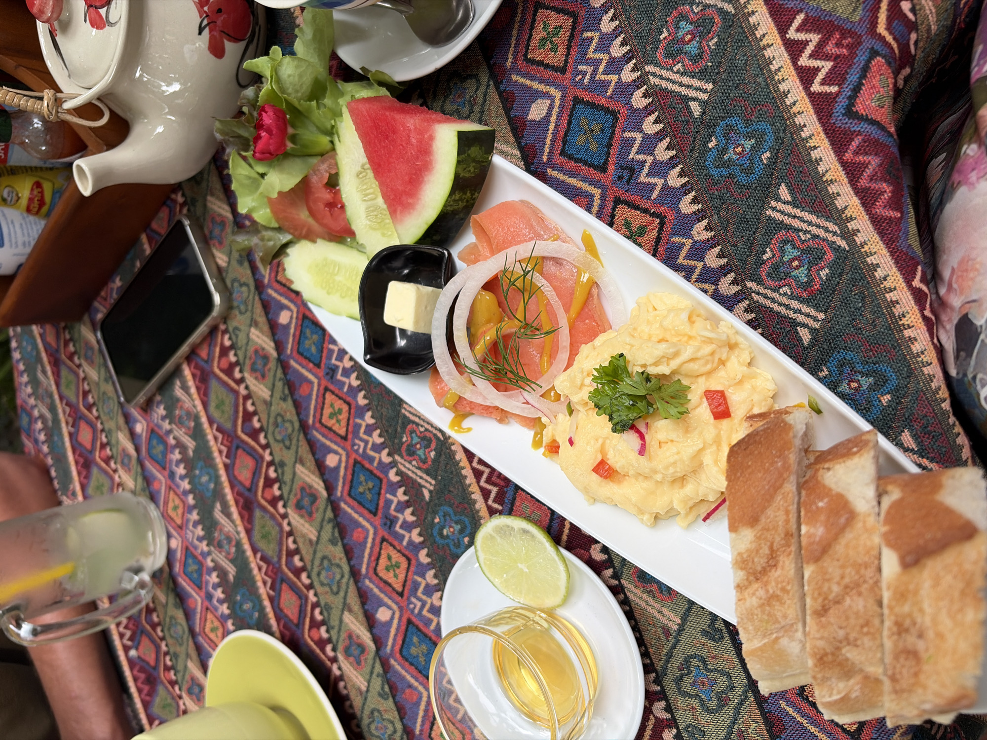 The image depicts a breakfast spread on a table, likely at an outdoor cafe or restaurant in Chiang Mai City Municipality, Thailand. A colorful, patterned tablecloth covers the surface. In the foreground, slices of toasted bread are stacked on a white plate. Behind the bread, a white rectangular platter holds scrambled eggs garnished with fresh green herbs and thinly sliced red onion and red bell pepper. To the left of the eggs, thinly sliced smoked salmon is arranged with thinly sliced red onion and dill, drizzled with mustard. A small dish of honey and a lime wedge sit to the left of the main platter. Further back, a salad of lettuce, cucumber slices, tomato slices, and a wedge of watermelon is visible. A small square of butter sits in a black dish. On the right side of the frame, part of a white teapot with a floral design and a white teacup with a spoon are visible. In the background, a glass mug filled with a pale green beverage and ice is present, along with a mobile phone. The overall scene suggests a relaxed breakfast setting.