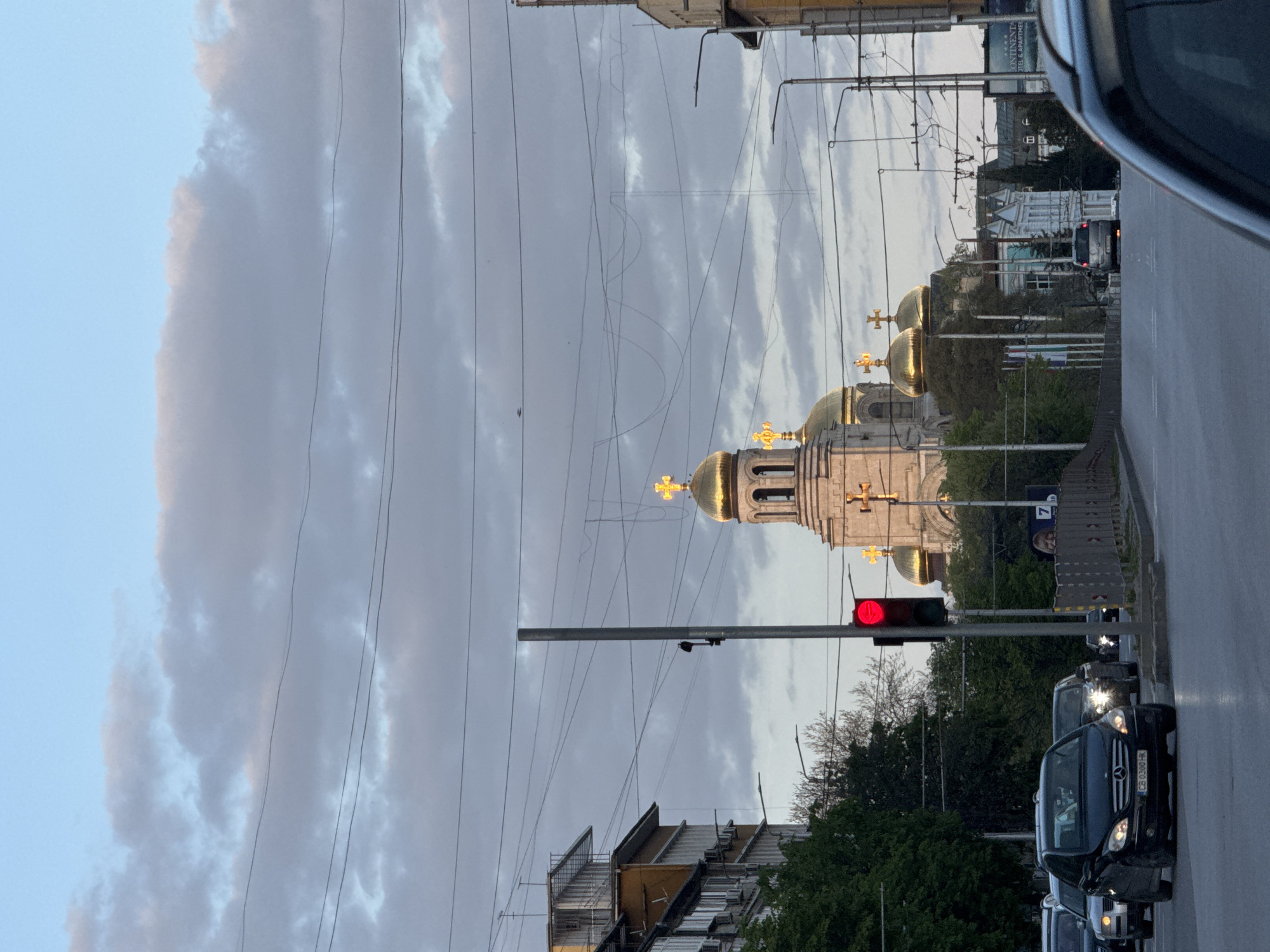 This is a portrait-oriented photograph depicting an urban street scene in Varna, Bulgaria, at dusk. The sky is partially cloudy with soft, diffused light, suggesting the transitional period between day and night.
In the mid-ground, several cars are visible, stopped on a multi-lane road, presumably at a red traffic light, which is prominently displayed on a pole. A dark Mercedes-Benz, bearing a Bulgarian license plate "CB 0300 HK", is visible in the foreground on the right side of the road.
To the left, a grand Orthodox church with multiple golden domes topped with illuminated crosses dominates the scene. The domes appear to be lit, enhancing their golden hue against the evening sky.
Along the street, various city buildings line both sides. On the right, a building features a partial sign that reads "CONTINENTAL OTEL & APARTME" (likely "Continental Hotel & Apartments"), and further down, a large billboard displays the number "7". Some buildings appear to have scaffolding or be under renovation. Trees and bushes provide greenery along the street. Numerous overhead utility wires crisscross the upper portion of the frame, adding to the urban feel. No specific individuals are discernible, and the overall mood is calm, typical of a city street during the evening commute.