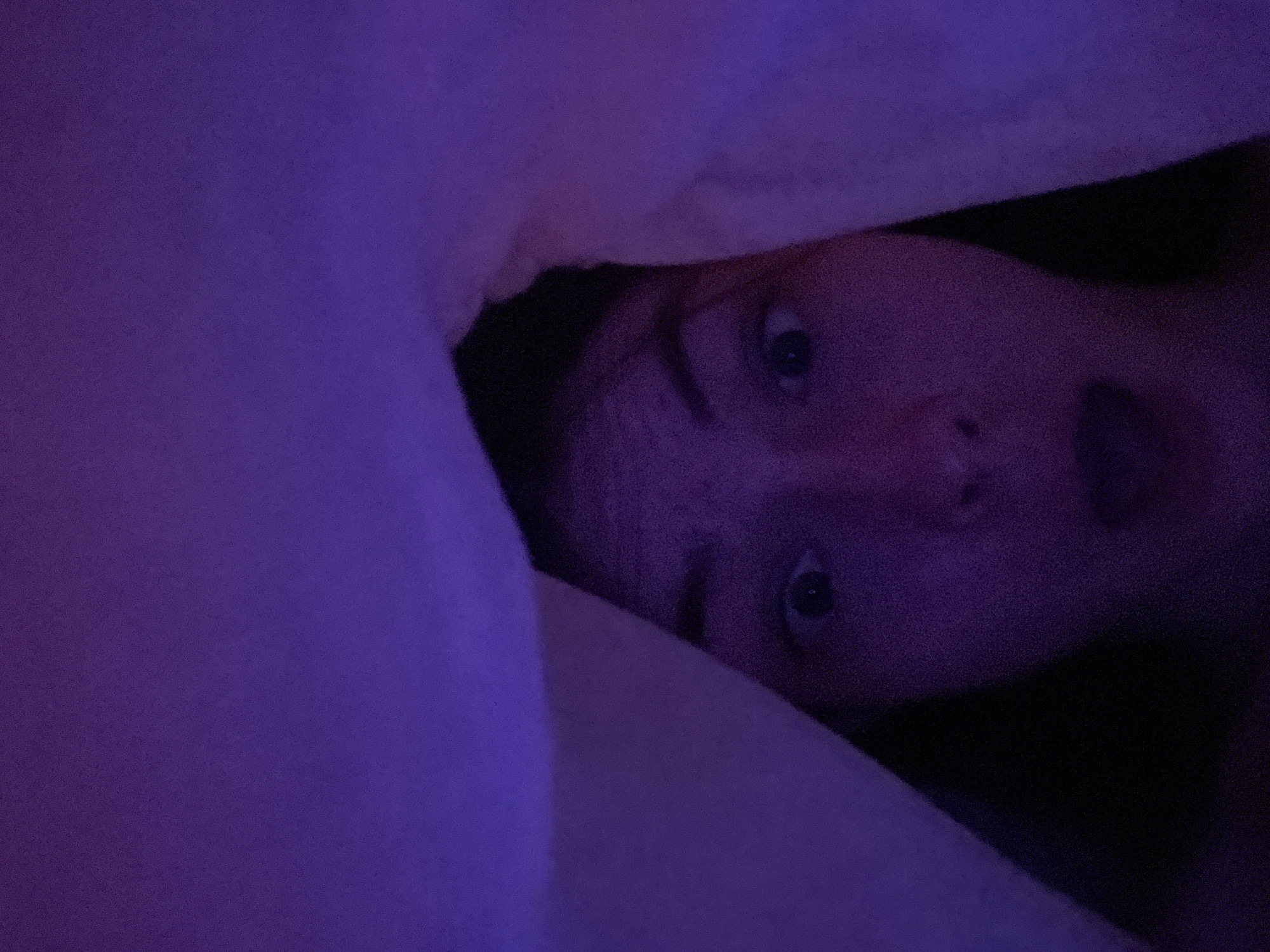 An indoor shot captured in low, purple-toned lighting shows a person's face partially emerging from under a dark purple or blue blanket/fabric. The camera angle is from above, looking down at the person who is lying on their back or side. Only the upper half of their face is clearly visible, with wide, startled eyes looking directly towards the camera. Their mouth is slightly open, contributing to an expression of surprise, shock, or perhaps playful alarm. The skin tone appears muted and slightly reddish-purple due to the ambient light. The surrounding fabric is a dominant feature, creating a sense of being enveloped or hidden. The scene suggests a nighttime setting or a very dark room illuminated only by an artificial purple light source. No specific location cues for Saint Petersburg, Russia are visible in the image itself. There is no visible text.