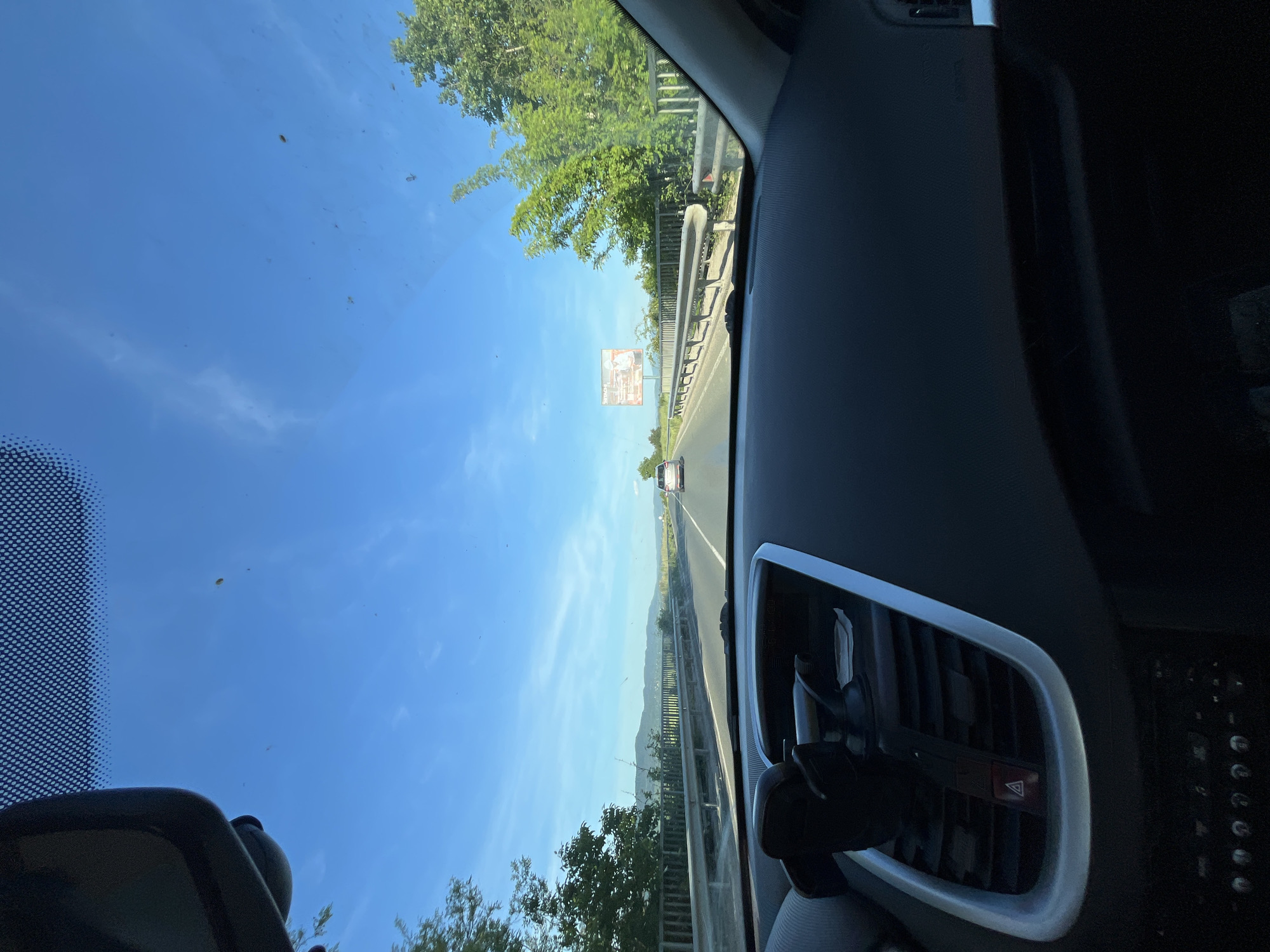 The image depicts a view from inside a car, looking through the windshield towards a highway. The sky is clear and blue, with some scattered clouds. There is an overpass with trees alongside the road. A billboard is visible in the distance. The interior of the car is visible, including the dashboard and the rearview mirror. The location is Unknown, Unknown.
