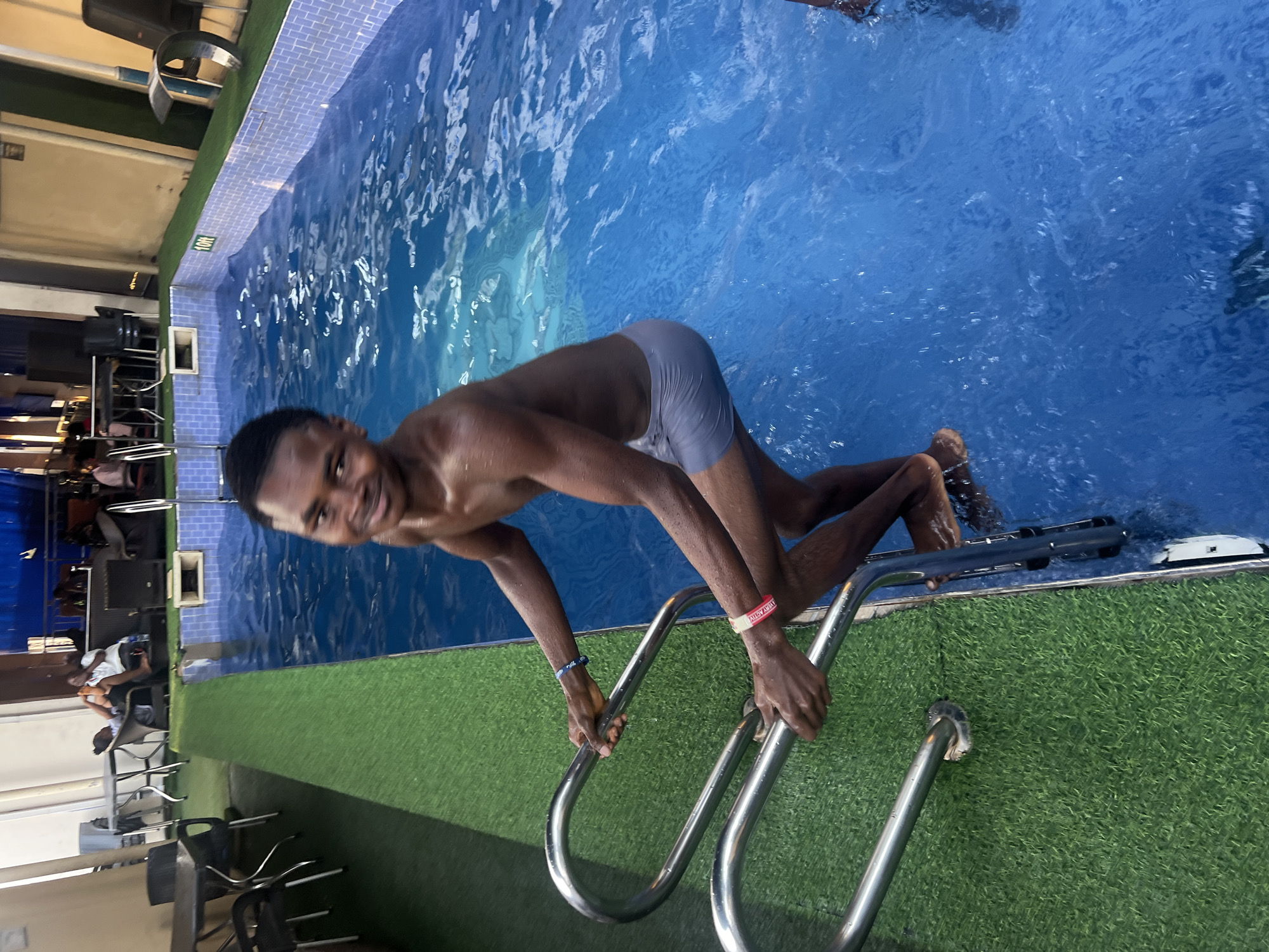 A young, smiling man, likely of Black African descent, is captured mid-action as he ascends a stainless steel ladder out of a swimming pool. He is wet and wearing light gray swim trunks. His hands grip the ladder railings, and he looks directly at the camera with a happy expression. On his left wrist, he wears a blue wristband, and on his right, a red/pink wristband with partially visible white text that appears to read "WHAT ACTIV."
The setting is an indoor or covered swimming pool area, likely a commercial leisure facility located in Nkpor, Nigeria. The pool features bright blue water and a tiled edge. Adjacent to the pool, artificial green turf covers the ground. In the background, tables and chairs are arranged, where several other individuals can be seen relaxing or socializing. Some large dark objects, possibly speakers or other equipment, are also visible. A white rectangular sign on the pool's edge displays "10M," likely indicating depth or length. The lighting suggests it is daytime, or a well-lit indoor space, and the overall atmosphere appears relaxed and recreational.
