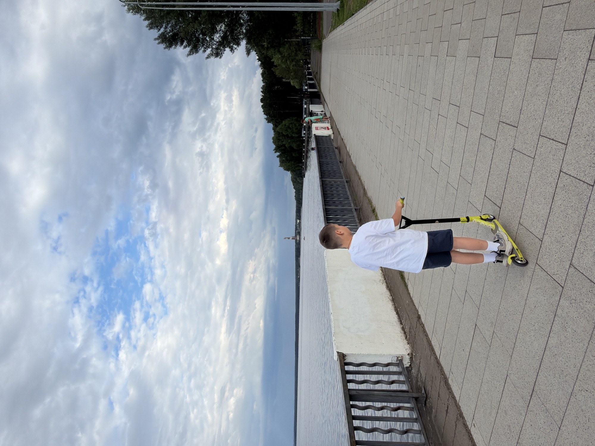 A boy rides a yellow scooter along a paved walkway next to a body of water. The boy, viewed from behind, wears a white t-shirt, dark shorts, white socks, and sneakers. The walkway is bordered by a low white wall and a dark metal fence on the left, beyond which is a wide expanse of water. The opposite side of the walkway is lined with trees and a tall street lamp. In the distance across the water, a lighthouse-like structure is visible, with a forested shoreline. The sky is cloudy with patches of blue. The scene is located on the embankment of the Votkinsk Reservoir in the city of Chaikovsky, Russia.