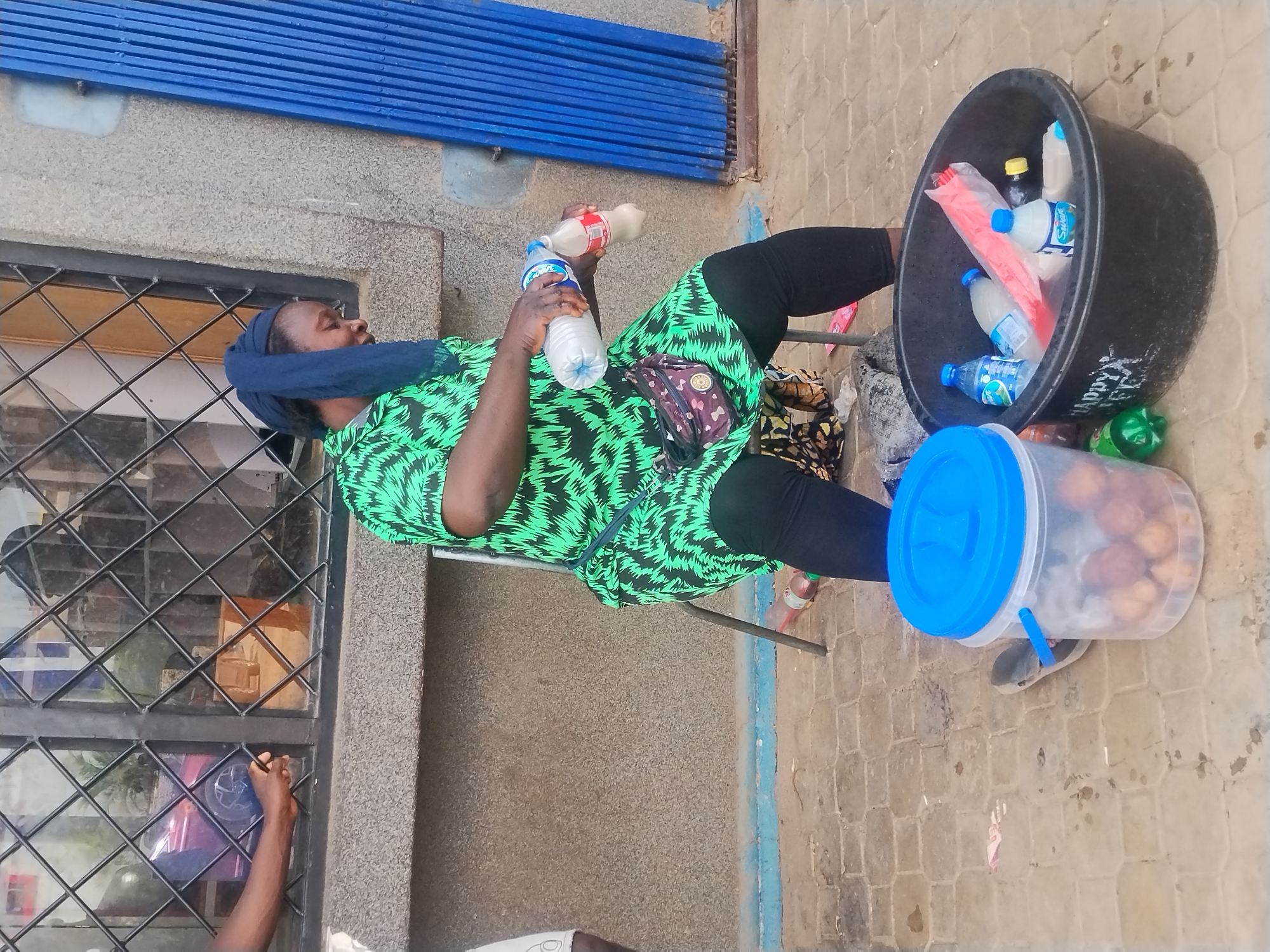A woman with a blue head covering, dressed in a green and black patterned top and black leggings, is seated outdoors holding two bottles of liquid, possibly water or juice. She has a waist bag around her midsection. Next to her is a large black tub filled with bottled drinks, some wrapped in a red plastic bag, and a clear plastic container with a blue lid, containing what appear to be round snacks or fruits. The scene is set on a stone-paved surface near a building. A window with a diamond-patterned grate is visible on the left. A person's arm is also visible near the window. The location appears to be a city, but the specific city is unknown.