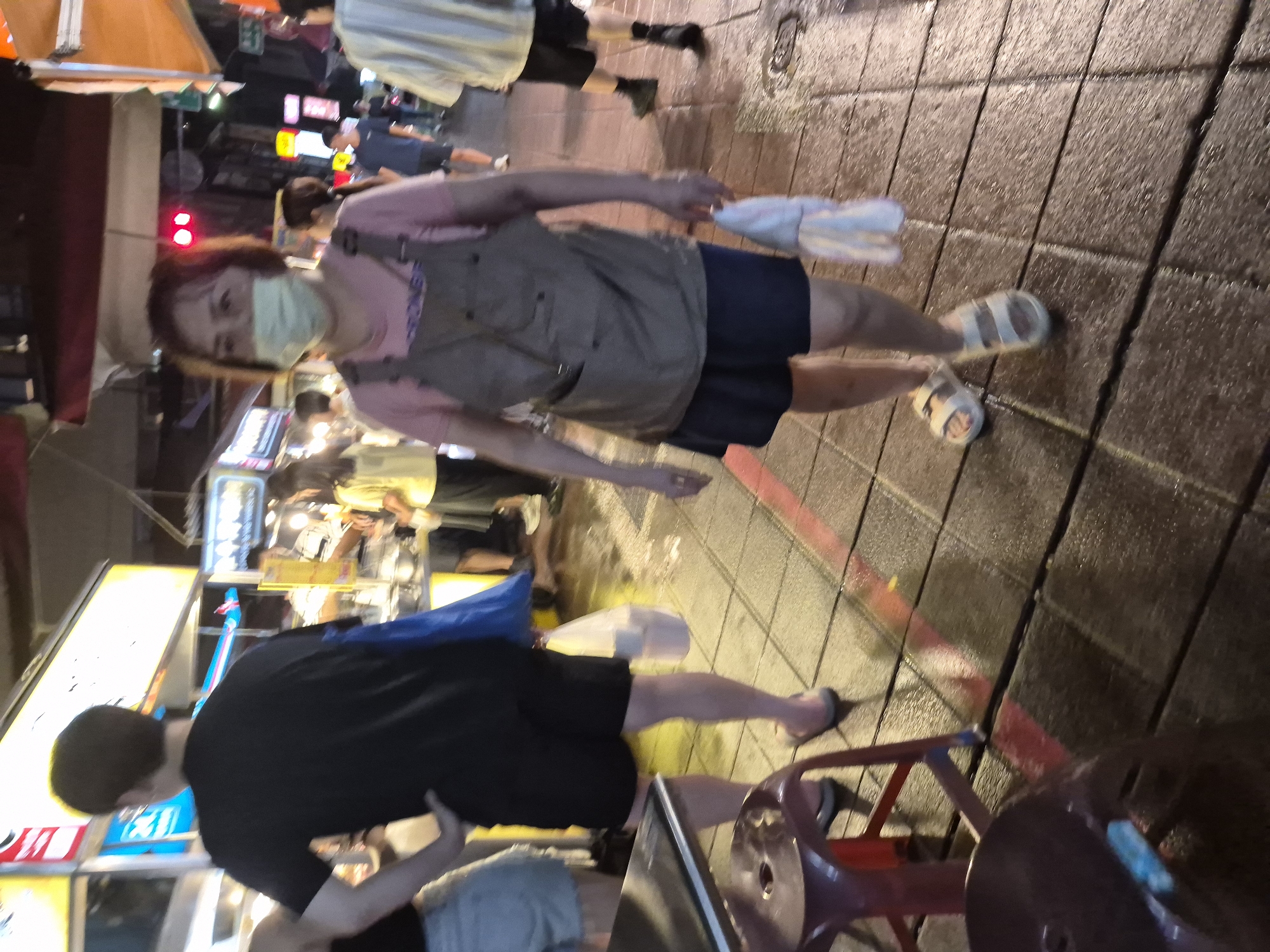 A woman wearing a light green face mask and a pink shirt with a grey apron is walking on a wet, tiled street at night. She is holding a white plastic bag in her right hand and is wearing white sandals. The street appears to be a market or a busy pedestrian area, with food stalls and neon signs visible in the background. The woman is walking away from the camera towards the right side of the frame. In the foreground, a red stool and a portion of a table are visible on the left. The overall scene suggests a bustling night market environment in Taipei, Taiwan, possibly after rain, given the wet surface of the street.