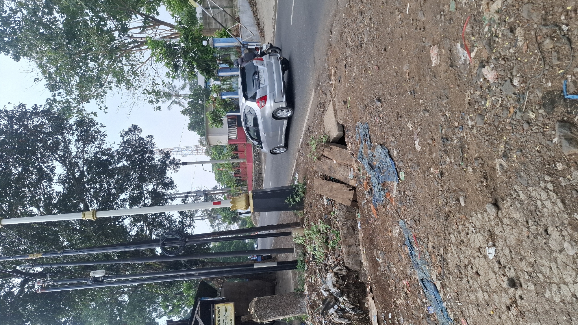 In Cilaku, Indonesia, a silver Honda Jazz is driving down a paved road. To the left of the road, there is a large pile of garbage and debris. The garbage includes plastic bags, wood planks, and various other discarded items. The area appears to be a roadside dump. In the background, there are trees, utility poles, and buildings. The sky is overcast.