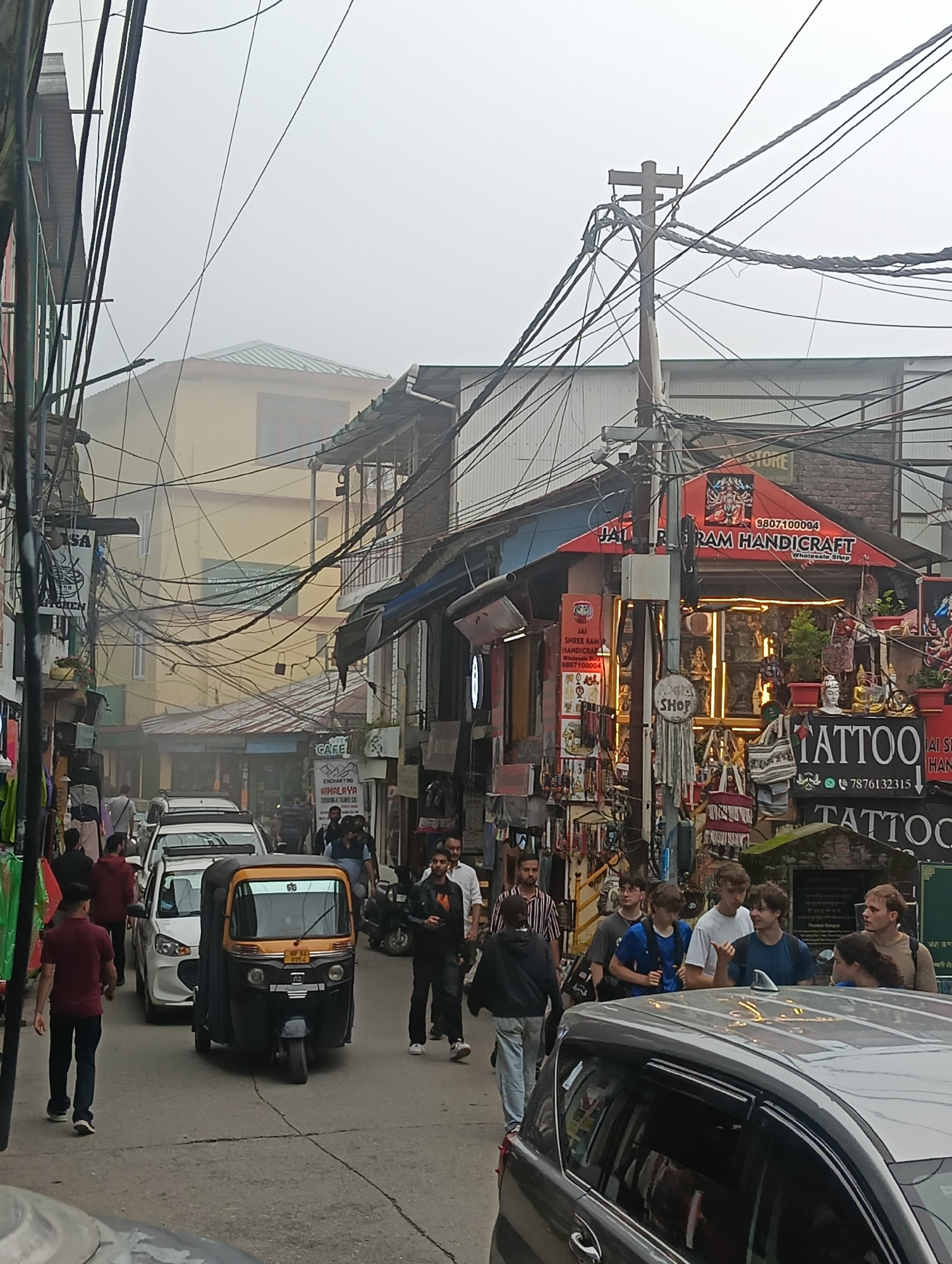 The image depicts a busy street scene in Dharamkot, India, characterized by a narrow road lined with shops and buildings. The atmosphere is hazy, suggesting either fog or pollution, with a plethora of overhead electrical wires creating a dense, tangled network.
In the foreground, an auto-rickshaw, primarily yellow and black, is positioned in the middle of the road, facing the viewer. To its left, a person in a maroon shirt and dark pants walks away from the camera. Several cars, including a white vehicle and parts of others, are visible on the road, indicating vehicular traffic.
The sidewalks are occupied by a mix of locals and tourists. On the right side of the street, a group of young men, seemingly tourists, are gathered. One individual, wearing a white t-shirt, is gesturing with his hand. Adjacent to them, a tattoo shop with prominent signage reading "TATTOO" is visible, displaying artwork and various products. Further down the street, more shops are seen, selling handicrafts, clothing, and other goods. A sign for "JAI RAM HANDICRAFT WHOLESALE SHOP" is prominent on the right side.
On the left side of the street, shops also line the road, with some displaying clothing and bags. Signs for "SAI KITCHEN" and "CAFE" are also present. People are seen walking in both directions, interacting with shops and navigating the crowded space. The buildings are multi-story, with balconies and awnings, and exhibit a mix of architectural styles. The overall impression is one of a bustling commercial area in a hilly region.