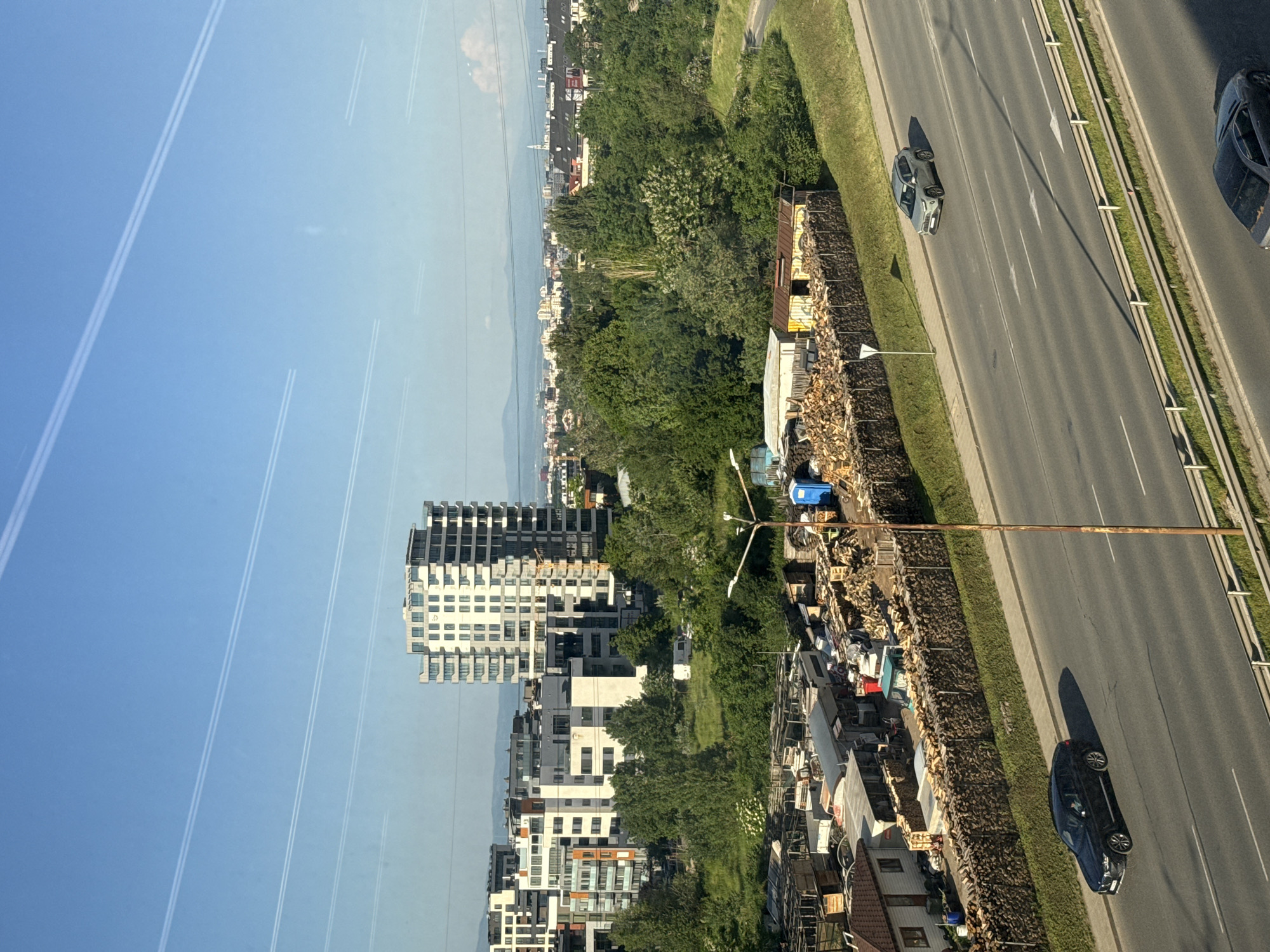 The image shows an aerial view of a city scene in Sofia, Bulgaria. Modern residential buildings are visible to the left, contrasting with a line of greenery separating them from a multi-lane road on the right. On the roadside, there is an assortment of discarded items and construction materials. Several cars are driving on the road. In the sky, there are contrails.