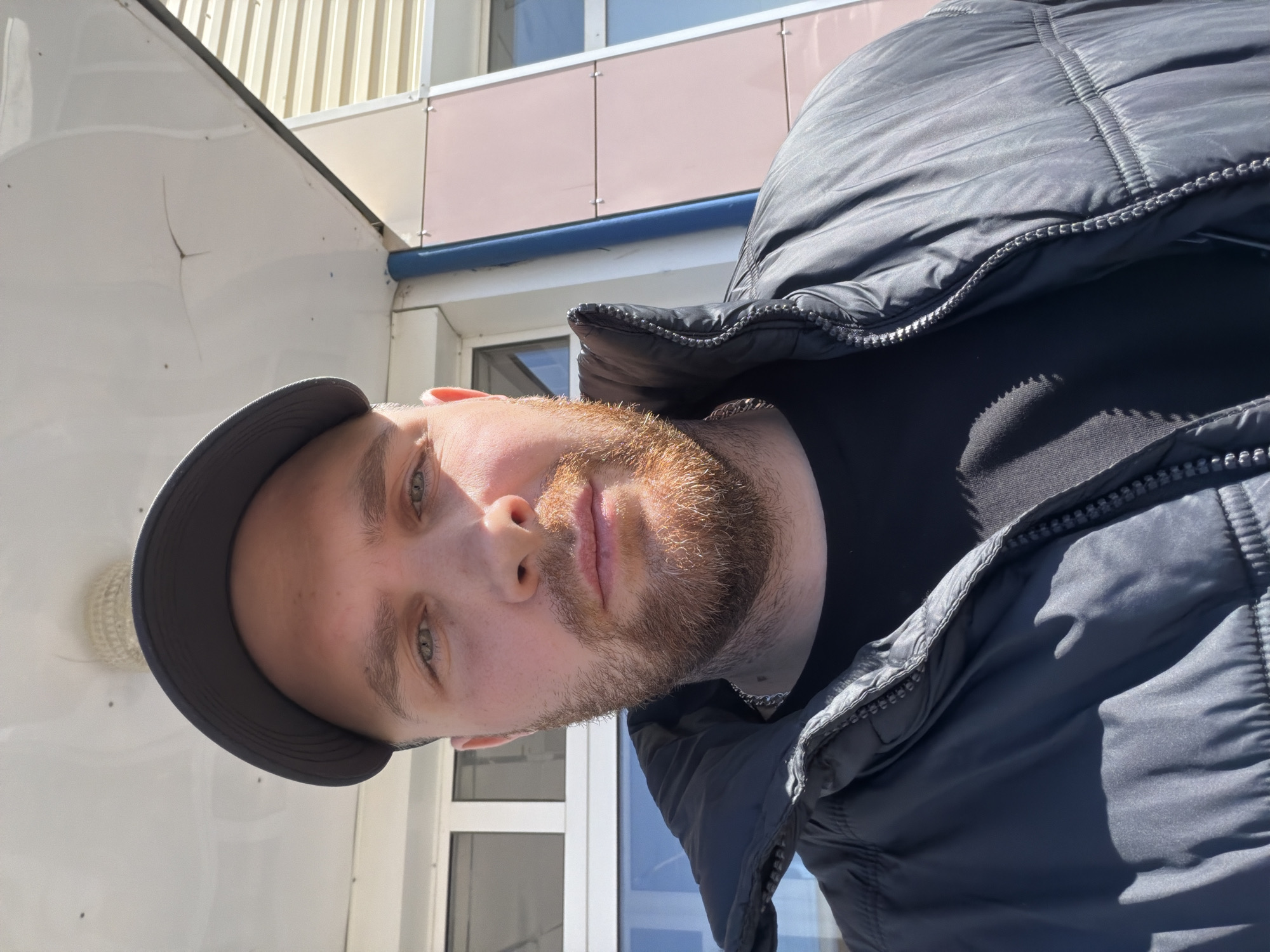 A medium-shot, close-up, portrait-style photograph shows a man wearing a dark-colored baseball cap and a dark, puffy jacket. He faces the camera. He has a light complexion, short blond hair, and a reddish-brown beard. Behind him is a light-colored building exterior with windows. The exact location in the city of Chaikovsky, Russia, cannot be determined from the image alone.