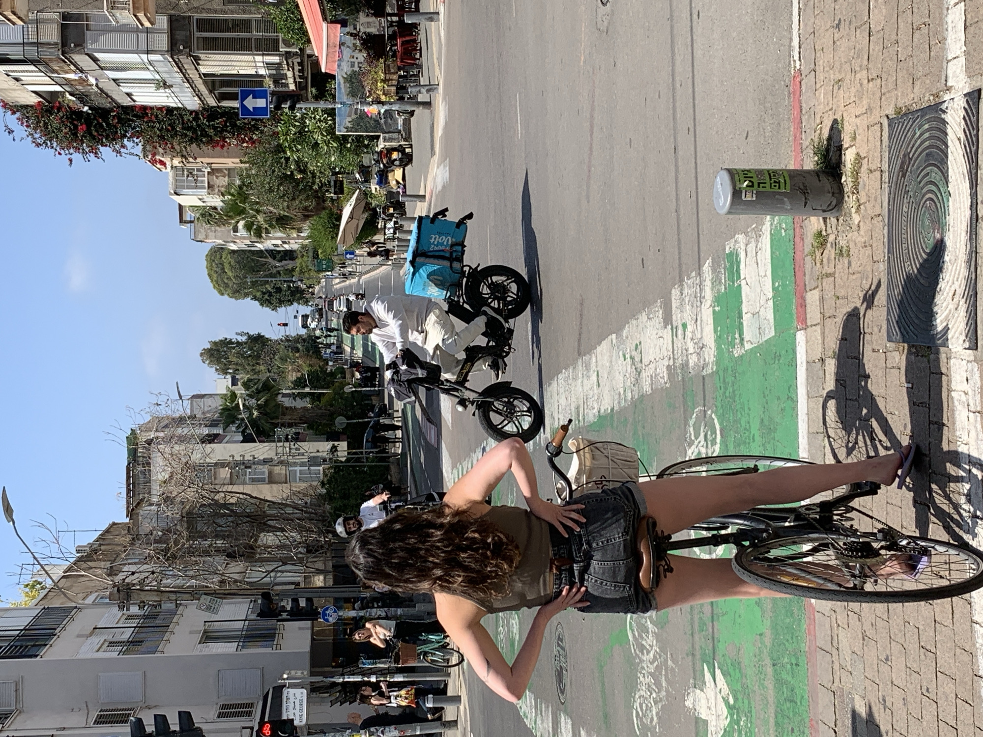 The scene is a street in Tel Aviv, Israel. A woman with long brown hair, wearing a brown top and denim shorts, is straddling a bicycle on a green bike path. A man is riding an electric scooter, wearing a white shirt and trousers. The scooter has a light blue delivery box attached. Buildings line the street, and there are traffic lights and street signs visible. A manhole cover is also visible in the corner of the image.