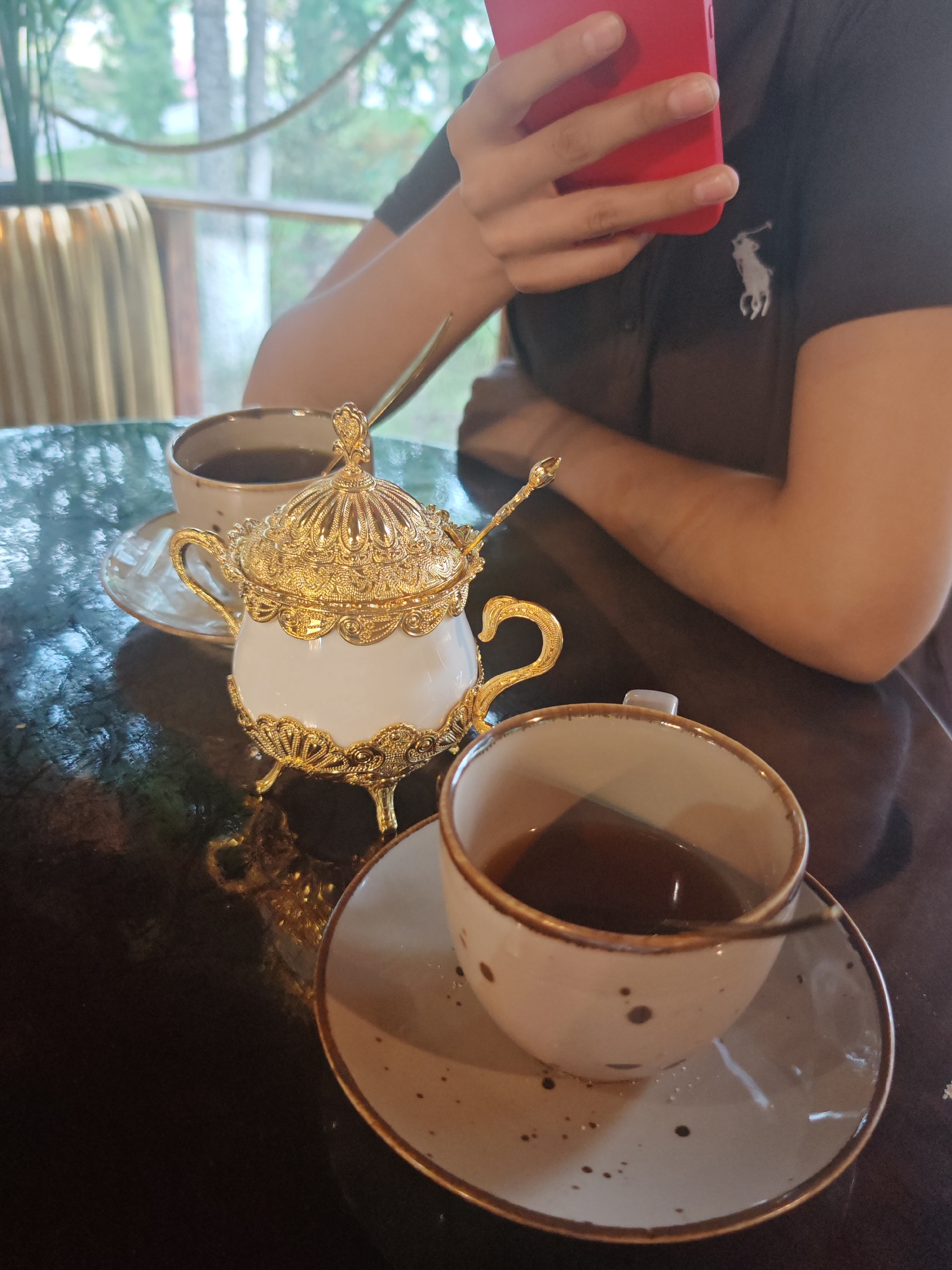 In a cafe in Novy Urengoy, Russia, a person sits at a dark, reflective table with two cups of coffee and a sugar bowl. The person, partially visible from the shoulders down, wears a dark polo shirt with a white horse logo. They hold a bright red smartphone in their left hand, suggesting they are engaged with their device.
The table is set with a white and gold ornate sugar bowl, complete with a small spoon. Next to it are two light-colored ceramic coffee cups with speckled patterns and a golden rim, each on its own saucer. One cup is in the foreground, its dark coffee reflecting the ambient light. The other cup is positioned behind the sugar bowl, also filled with coffee.
The background suggests an indoor setting with a view of trees and greenery through a window or glass partition, hinting at an outdoor natural environment. The lighting appears to be natural, possibly from daylight filtering in. The overall scene evokes a casual and relaxed atmosphere, perhaps a moment of personal downtime during the day. There is no visible text in the image.