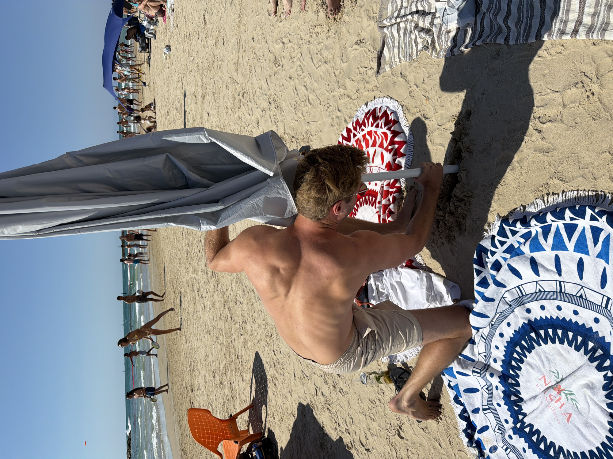 The image depicts a man on a beach, seemingly setting up a beach umbrella. He has a fair complexion, short blonde hair and is shirtless, wearing light-colored shorts.
The beach appears crowded with people near the water's edge, suggesting a popular location. People can be seen walking and playing in the water, with clear skies above.
Several beach towels are spread out on the sand around the man, including one with red and white patterns and another with a blue and white design. A small orange plastic chair is visible near his feet, as well as a dark object, possibly a bag. There is a glass on the sand.
Given the beach setting and general context, it is likely this image was taken in Tel Aviv, Israel, known for its Mediterranean coastline and beach culture.