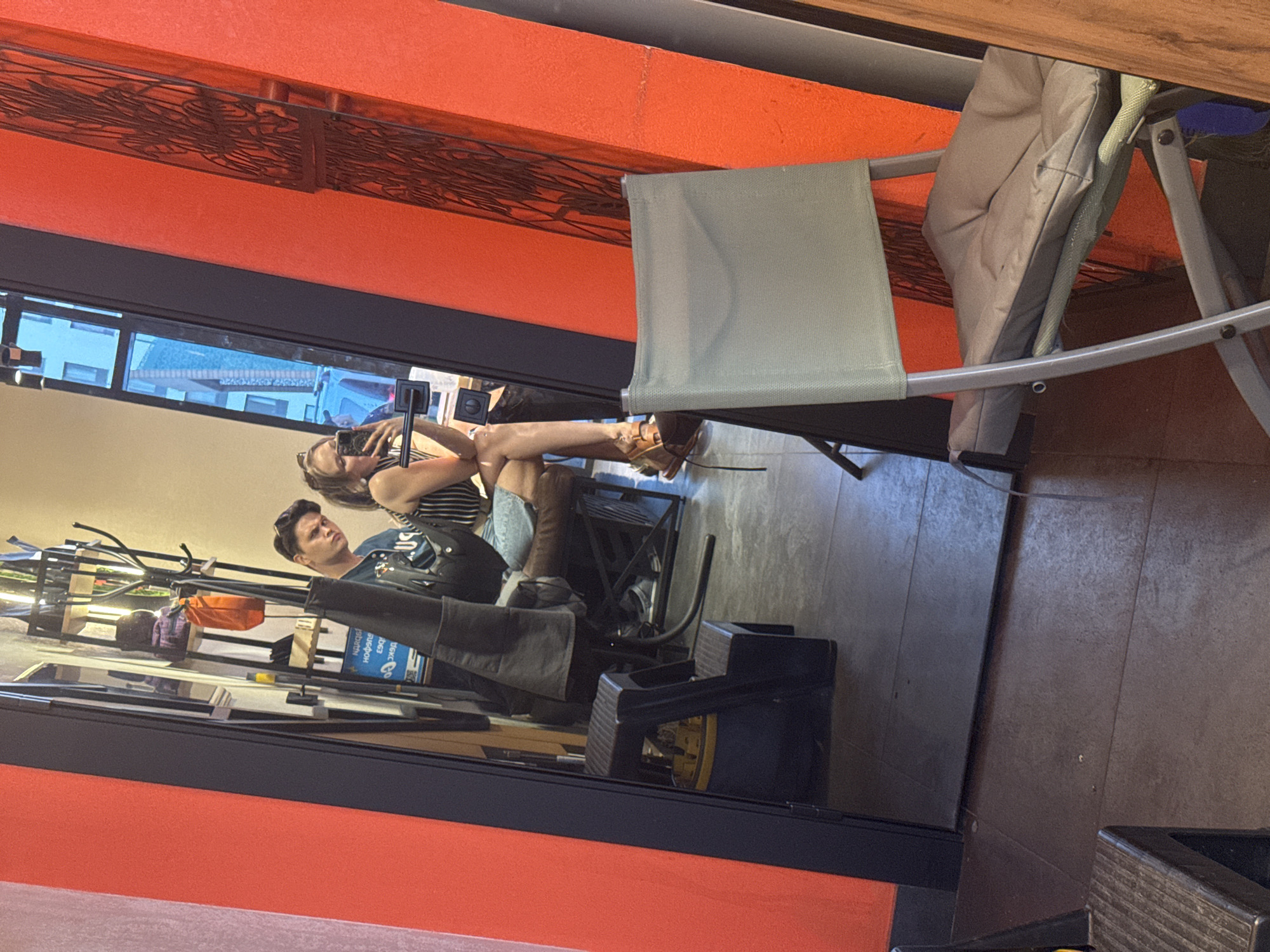The image shows a reflection in a window. In the reflection, a woman is taking a picture of herself and a man who is seated next to her. The woman is wearing a striped top, denim shorts, and brown boots. The man is wearing a blue shirt. In the background of the reflection, there is a rack with clothing and other items. Outside the window, there are buildings. The location of the reflection is likely in Izhevsk, Russia, based on signage that is not visible in the original image but is present in the crops. In the foreground, there is a metal folding chair and a wooden table. The area around the window is painted orange.