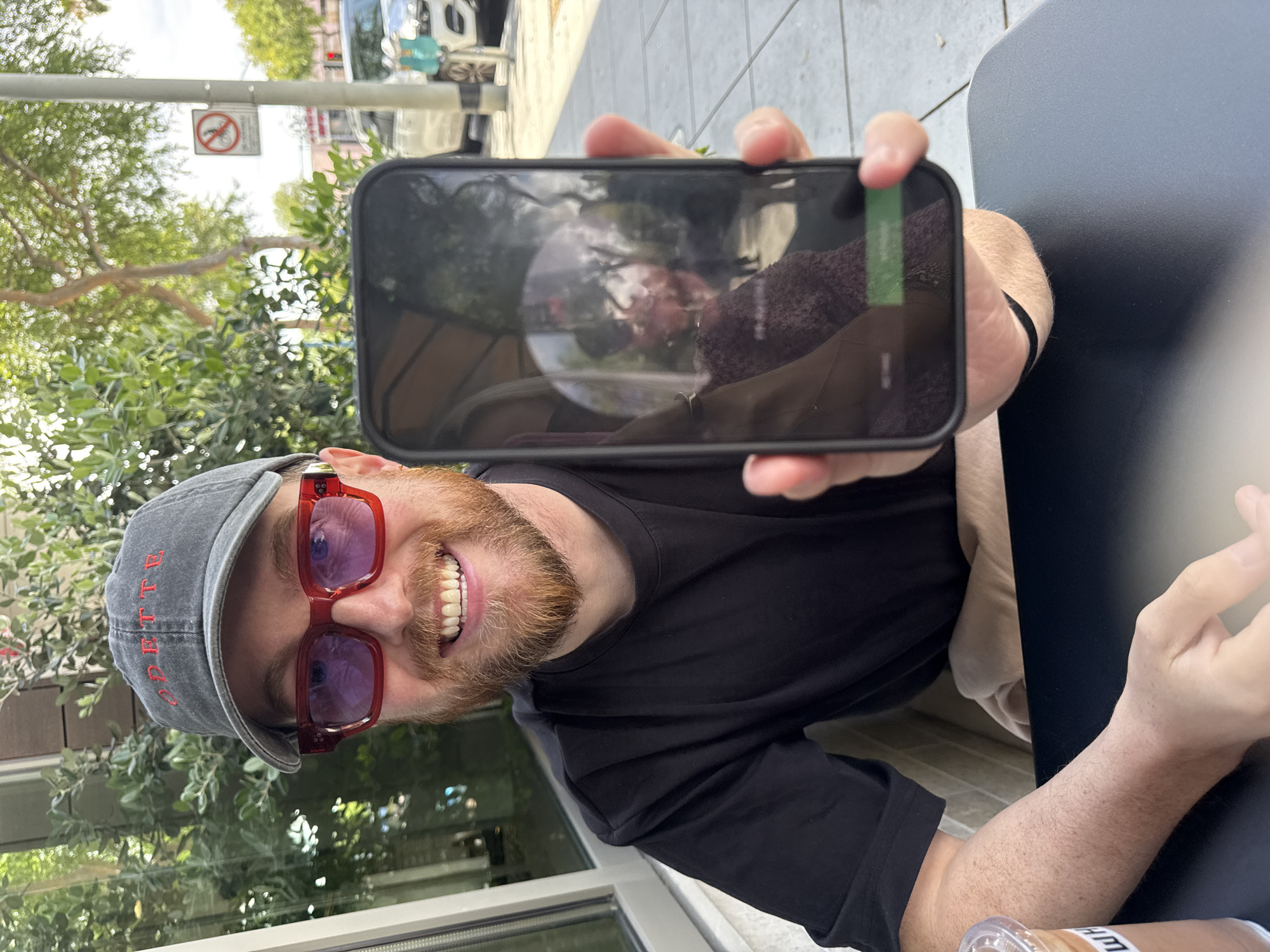 The photograph captures a smiling man with a red beard and red-tinted sunglasses, wearing a gray baseball cap and a black long-sleeved shirt. He is holding up a smartphone, displaying a reflection that seems to include foliage, a white object, and a person. The background includes a street scene, possibly in West Hollywood, indicated by street signs and trees. The subject appears to be sitting at an outdoor table, with a dark surface visible to the right and a glimpse of what might be a beverage container in the lower right corner. The overall scene suggests an outdoor cafe setting.