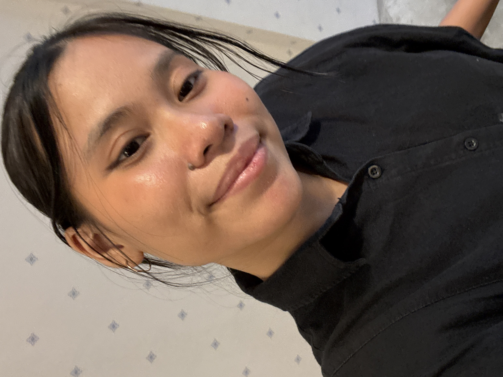 A medium shot photograph shows a young woman with dark hair and light skin, wearing a black collared shirt. She is looking at the camera and smiling slightly. Her hair is pulled back, and she has a small hoop earring in her left ear. The background is a wall with a subtle diamond pattern. The lighting appears to be natural or from a soft artificial source, highlighting the sheen on her skin. The image is cropped to focus on the woman's face and upper torso. The scene does not provide any specific details that would allow for a location to be identified in South Jakarta, Indonesia.