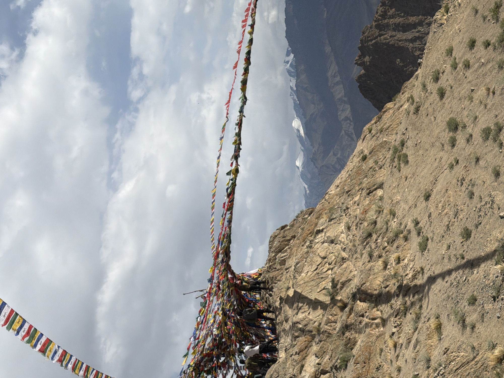 This image depicts a mountainous landscape, likely in the vicinity of Leh, India, characterized by prayer flags strung across the terrain. In the foreground, a rocky, sparsely vegetated slope dominates the lower portion of the frame. The texture of the rock is rough and varied, with small shrubs and patches of dry grass scattered across its surface. A long, dark shadow, likely cast by a flagpole or structure, stretches diagonally across this slope.
Further up, a cluster of individuals, appearing to be tourists or locals, stand on a rocky outcrop. They are casually dressed and some are looking towards the prayer flags, suggesting they are observing or participating in a ritualistic activity. The prayer flags are colorful and numerous, strung in long lines and cascading from what appears to be a central point, creating a vibrant display against the cloudy sky. The flags are made of cloth and are fluttering in the wind, adding a sense of movement to the scene.
In the background, a vast expanse of mountains stretches towards the horizon. These mountains exhibit a rugged, layered appearance, with some peaks capped with snow. The sky is overcast with a mix of white and grey clouds, hinting at potentially changeable weather conditions. The overall impression is one of a remote, spiritual location, emphasizing the natural beauty of the mountainous region and the cultural significance of the prayer flags.
