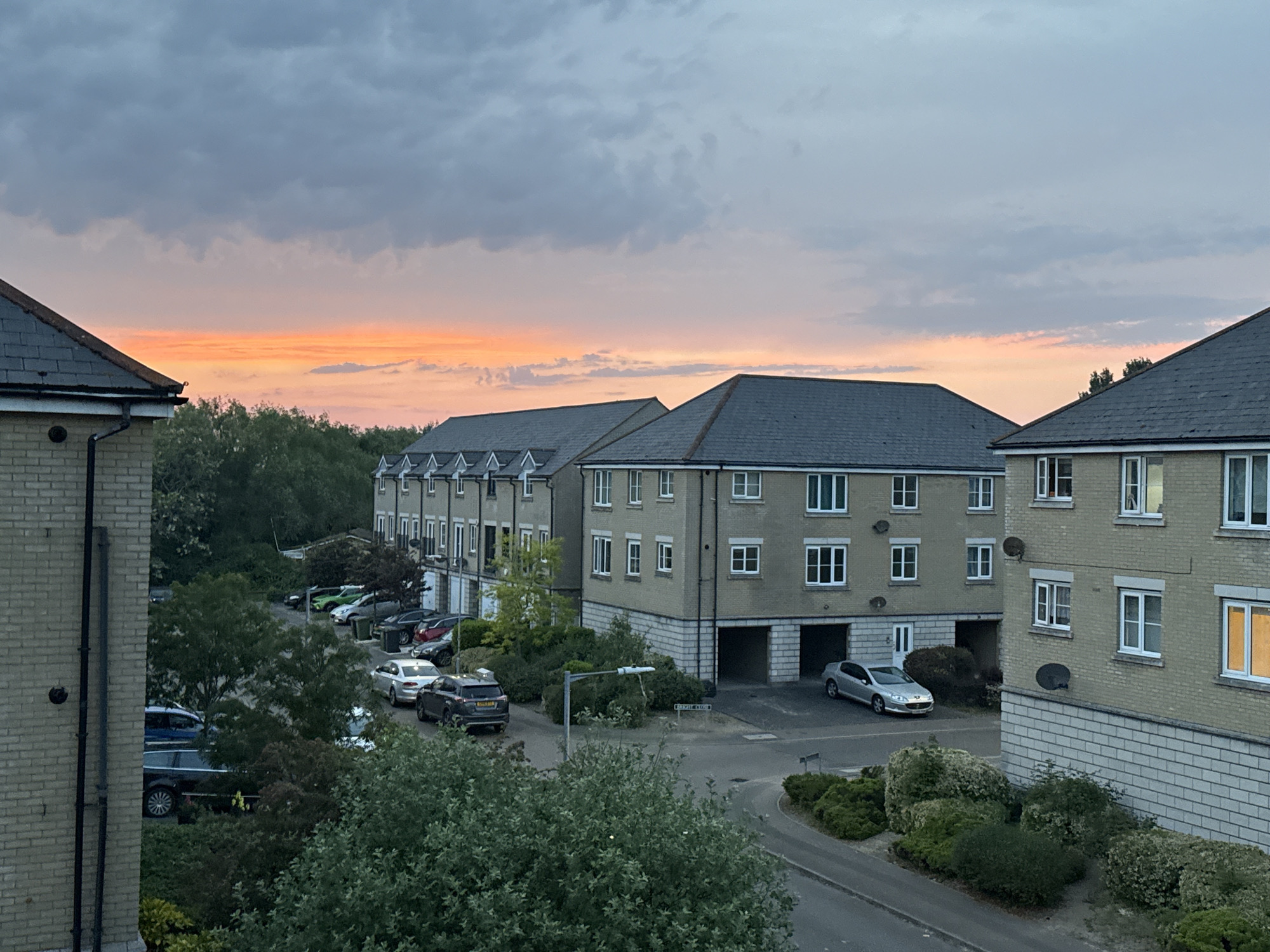 An elevated view shows residential buildings in Great Yarmouth, United Kingdom, at sunset. Three-story brick apartment buildings with gray pitched roofs frame the view. A row of parked cars lines a street alongside the leftmost building. Trees are visible between the buildings. A street lamp stands on a street corner. The sky is filled with clouds, tinted orange by the setting sun.