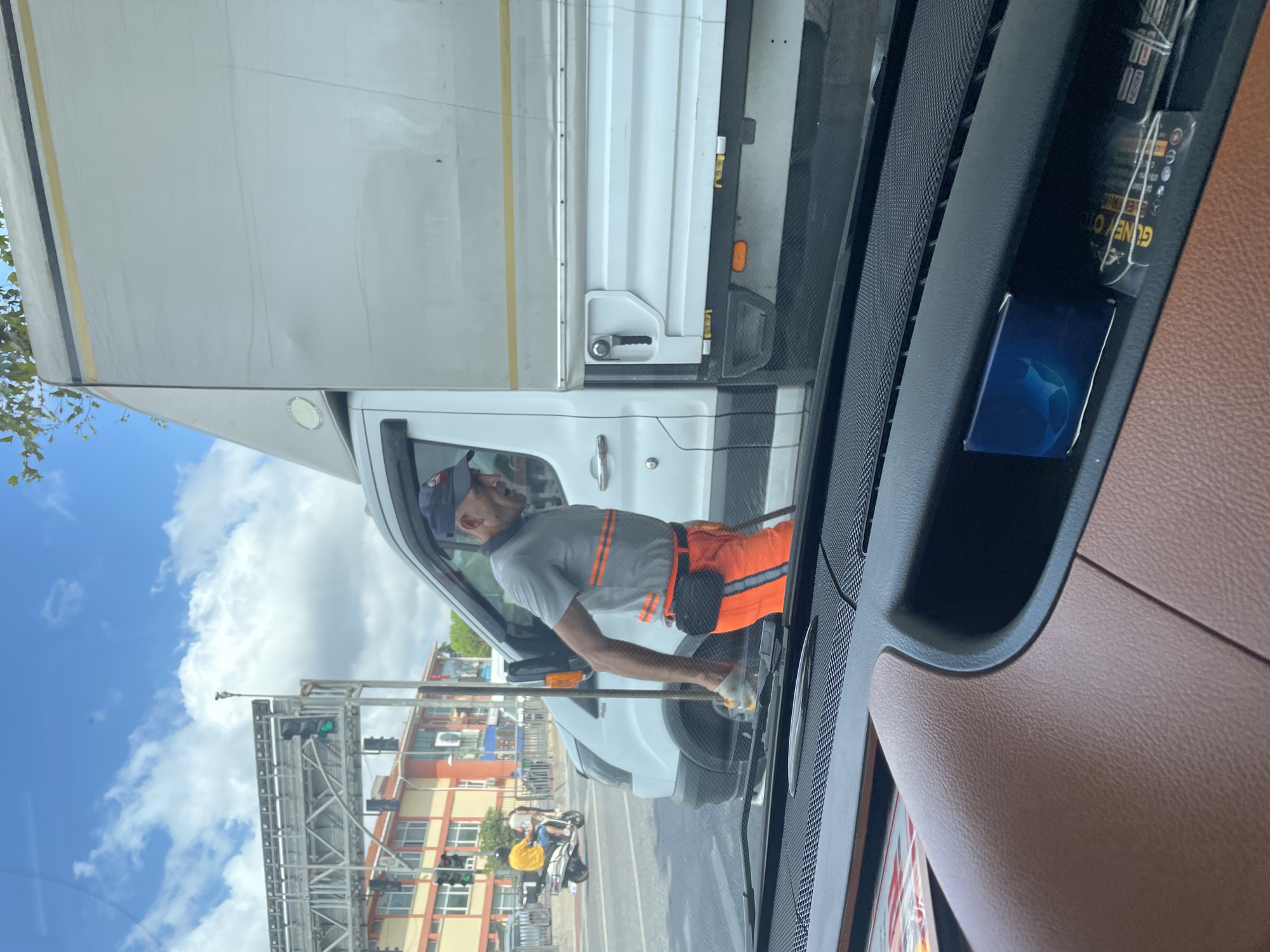 A man in an orange uniform with reflective stripes is standing next to a white truck. He is holding a pole and appears to be performing some kind of work. The scene is in a city with traffic lights and buildings in the background. The image is taken from inside a vehicle looking out at the street.