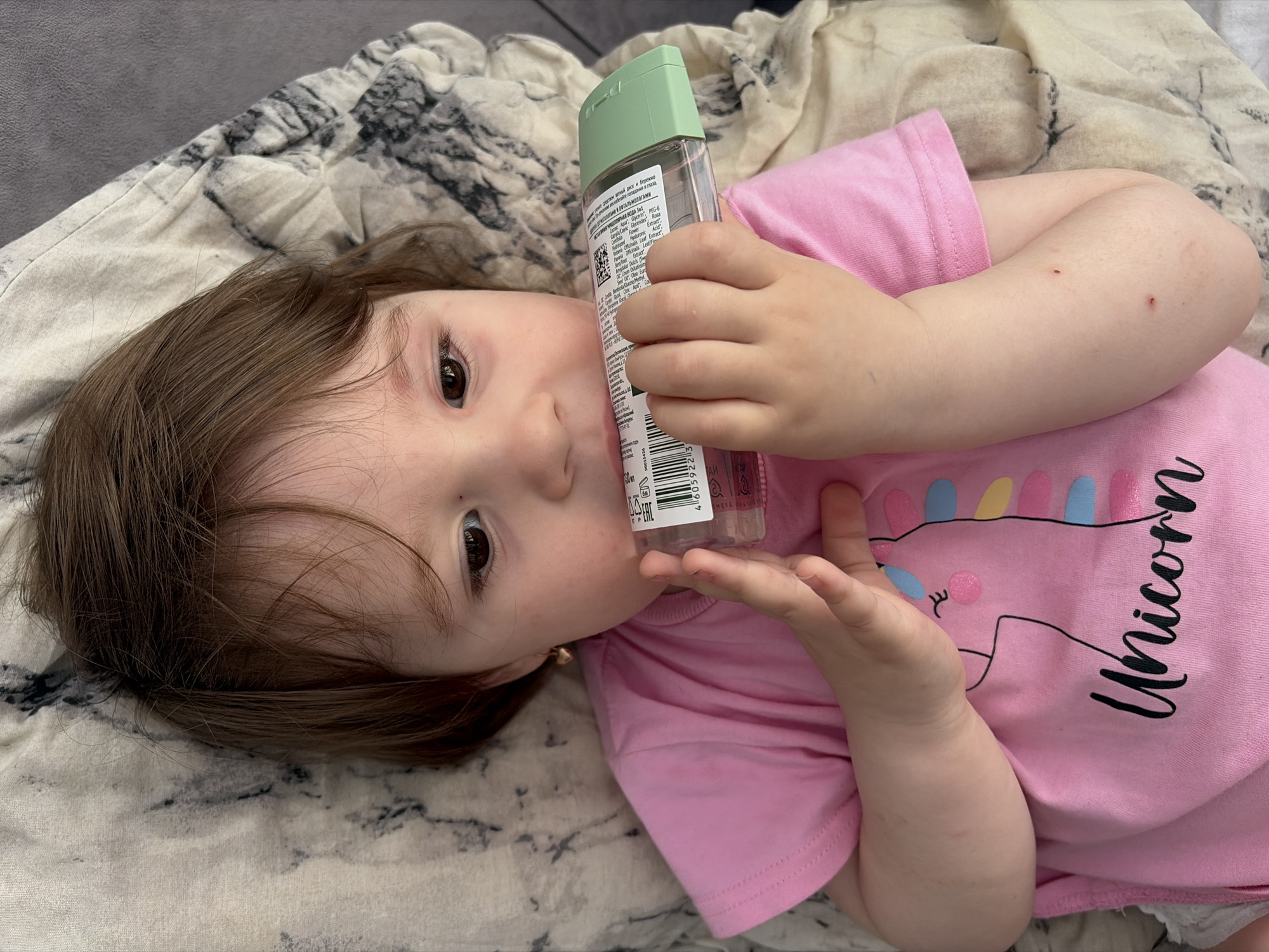 A young child with brown hair and dark eyes is depicted lying on a patterned blanket, looking directly at the viewer. The child is wearing a pink short-sleeved t-shirt featuring a cartoon unicorn with a colorful mane and the word "Unicorn" written next to it. The child's left arm is bent, and they are holding a clear bottle with a light green flip-top cap. The bottle's label, which has text in a foreign language (likely Russian, given the location context), is visible. Small, reddish marks are noticeable on the child's right arm. A small, golden earring is visible in the child's left ear. The child's expression is neutral and curious. The background consists of a light-colored blanket or fabric with a dark, abstract, marbling pattern.
Visible text includes:
* On the t-shirt: "Unicorn"
* On the bottle label: "EAC", a barcode starting with "46059223", "MICELARNAYA VODA" (Micellar Water), and partial ingredient listings like "Capric Glycerides," "Rosa Leaf Extract," "Olea Eur." There's also some text that appears to be instructions or warnings, partially reading "ОЙ диск и бережно" (cotton pad and gently) and "ния в глаза" (into eyes). "ТАЙ" is also partially visible.
The scene appears to be indoors, with the child casually resting on a surface. The setting is Chaikovsky, Russia.